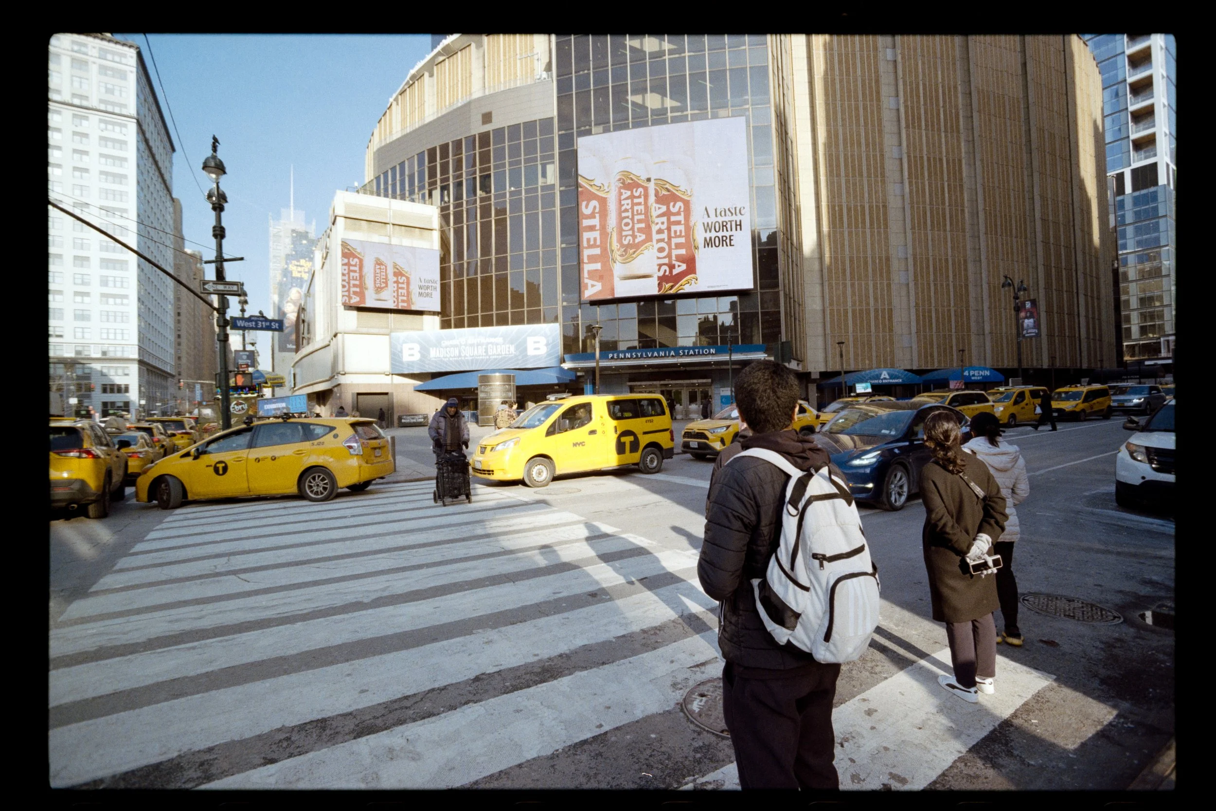 People waiting to cross a busy city intersection near Pennsylvania Station with yellow taxis and tall buildings, billboards, and blue sky