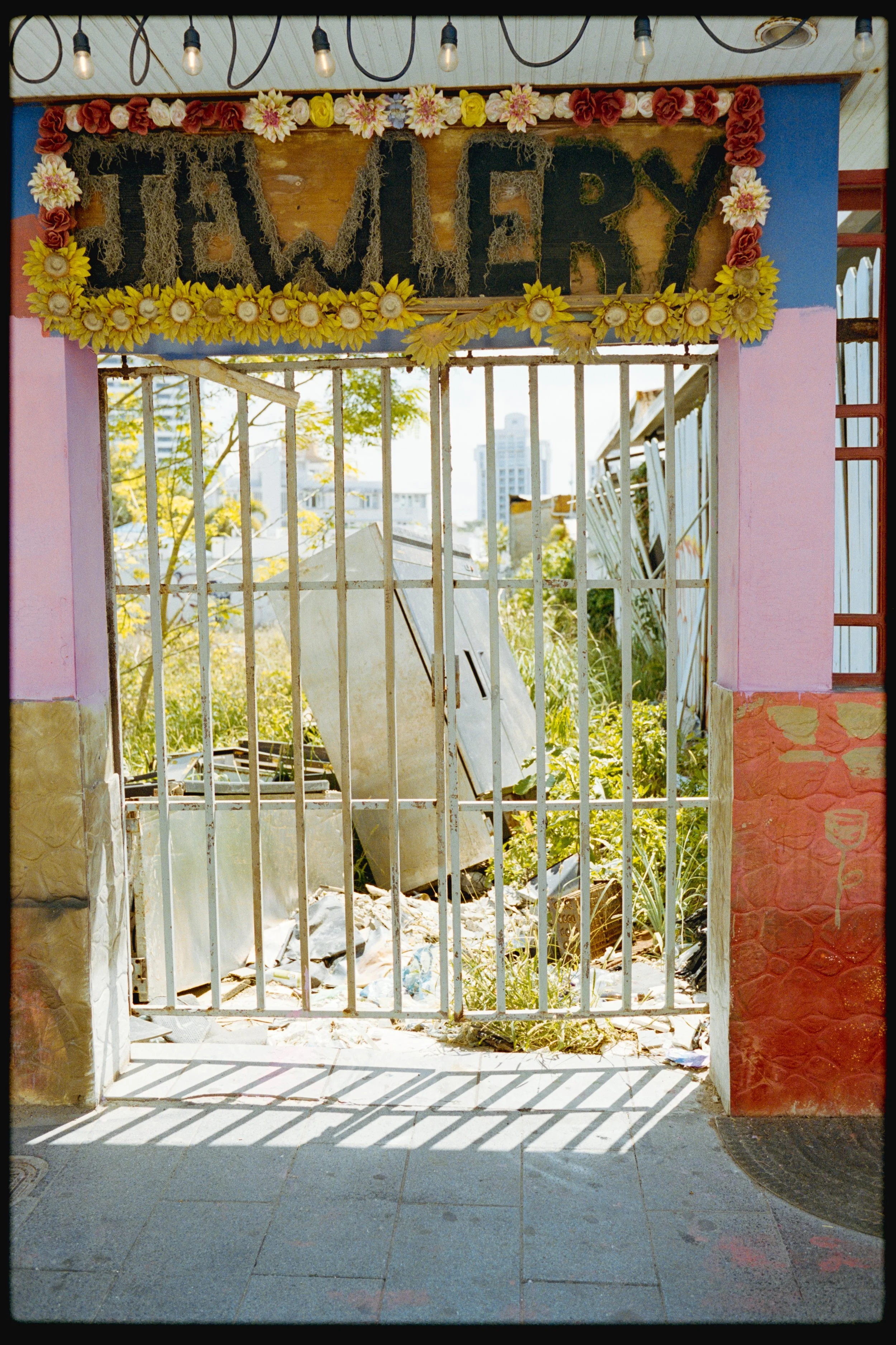 A decorative sign reading 'FARMERY' hangs above a white metal gate. The sign is adorned with flowers and moss, casting shadows on the tiled floor below. Behind the gate, an overgrown area with scattered debris and trees is visible, with urban buildin