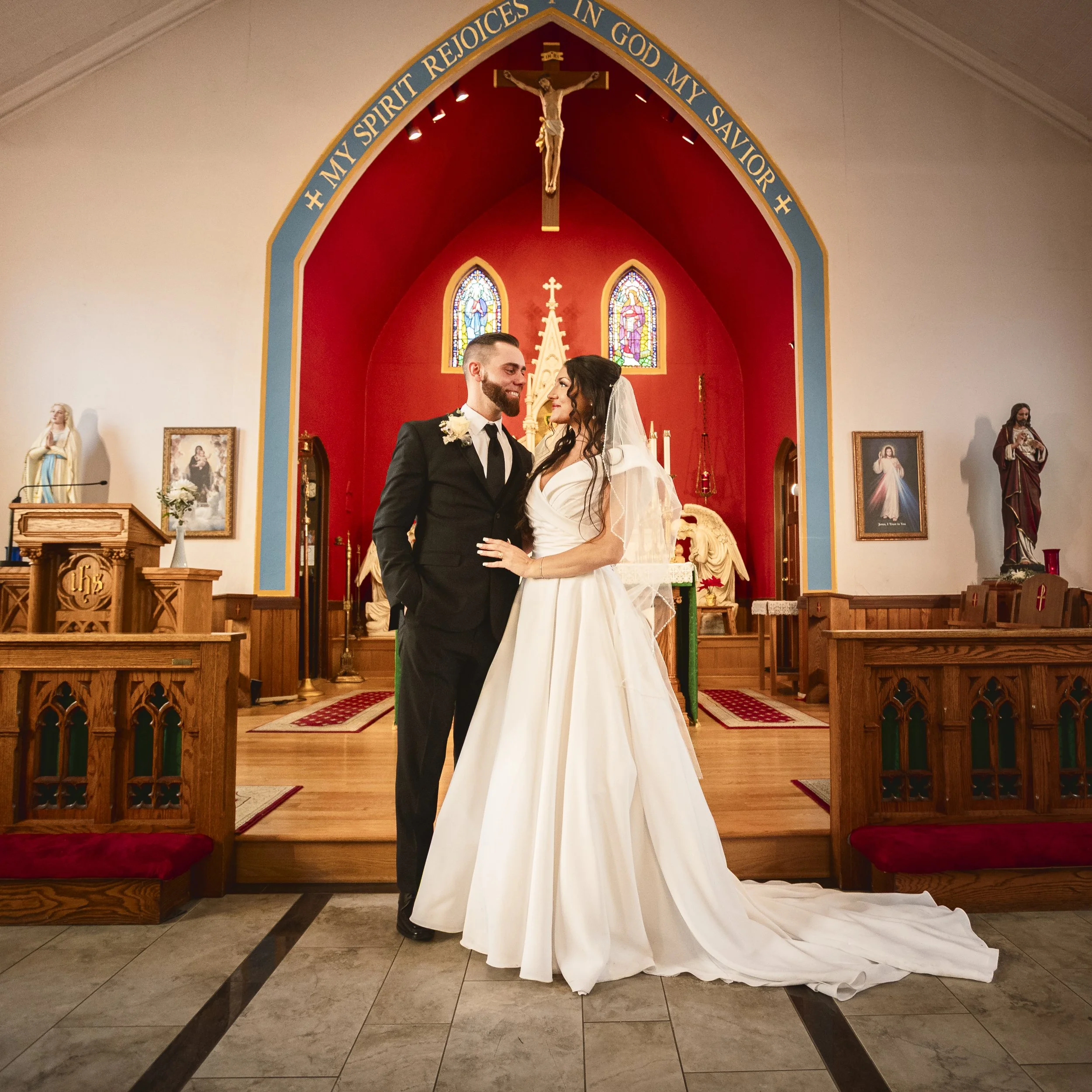 A bride and groom standing together inside a church, facing each other and smiling. The bride is wearing a white wedding gown and veil, and the groom is dressed in a black suit and tie. The church interior features religious statues, stained glass wi