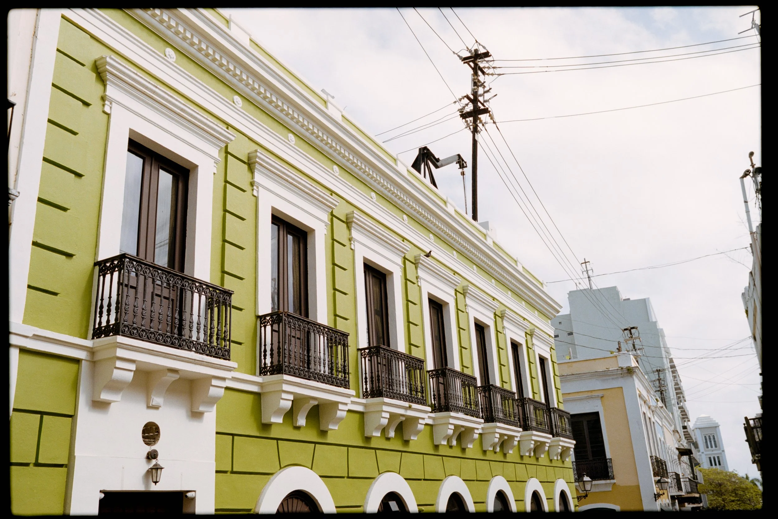 A colorful building with green and white exterior, featuring multiple windows with decorative black wrought-iron balconies and white arches at the ground level. Overhead, wires and utility poles are visible against a partly cloudy sky.