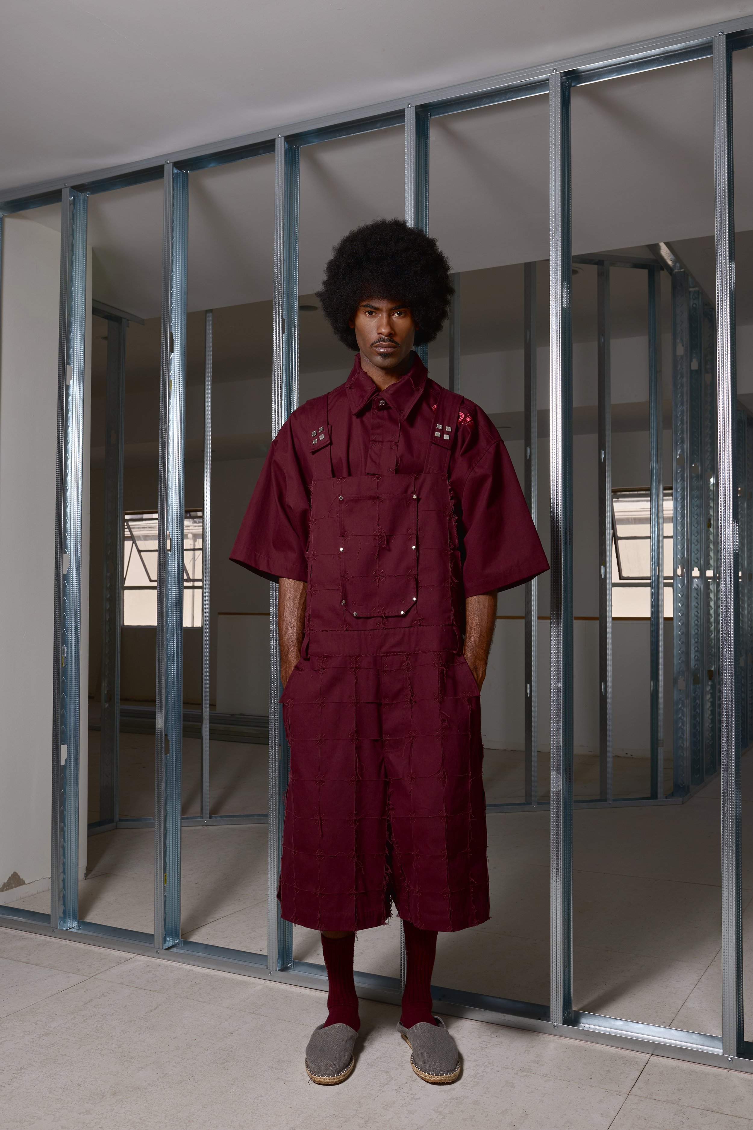 A man with an afro hairstyle standing in front of metal framing in an unfinished room, wearing a maroon outfit with a sleeveless dress over a shirt, maroon socks, and gray slip-on shoes.