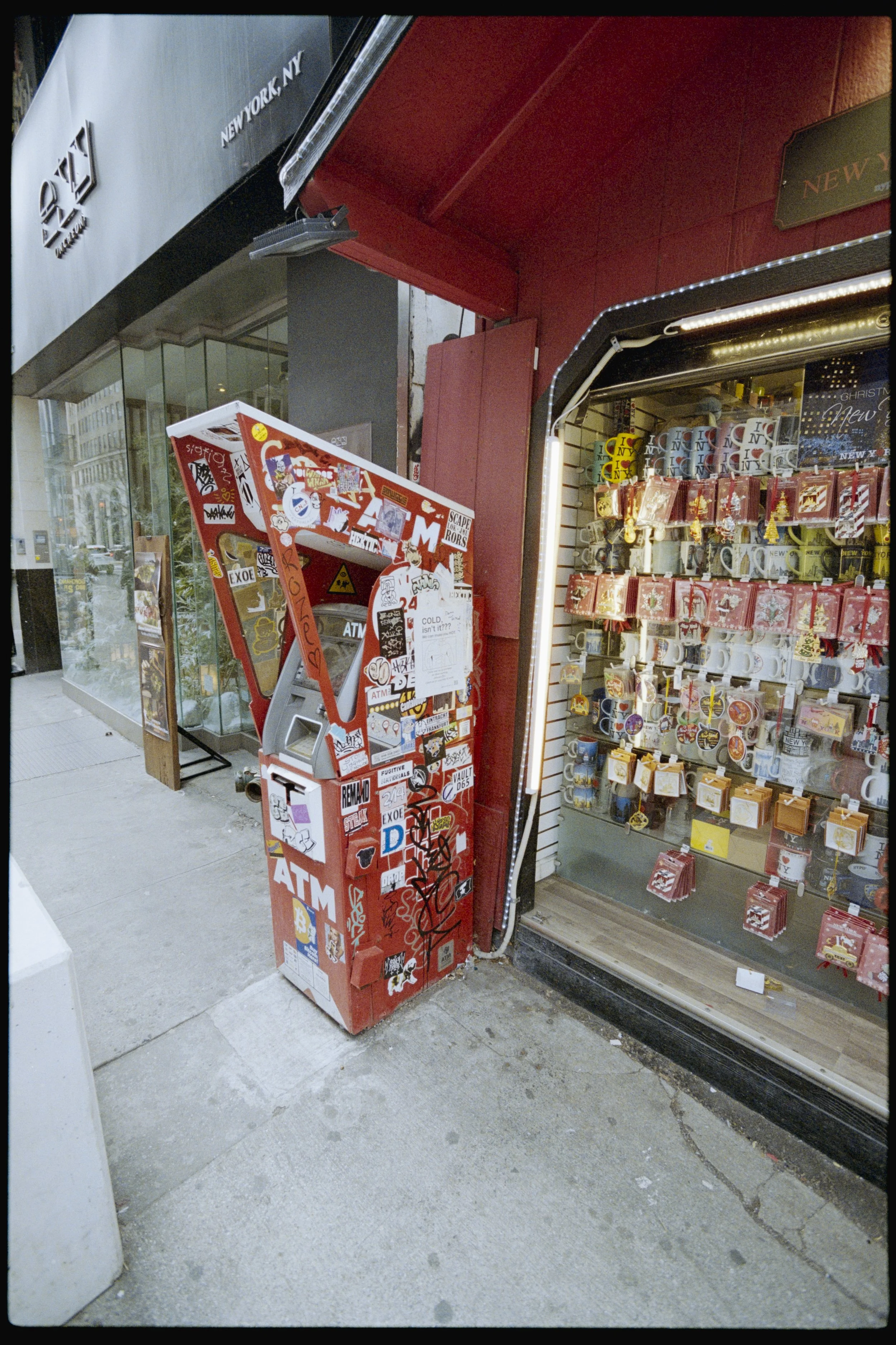 Street view featuring an ATM machine covered with stickers and graffiti positioned outside a shop that sells mugs with various designs. Reflections of buildings and trees can be seen in the shop's glass window.