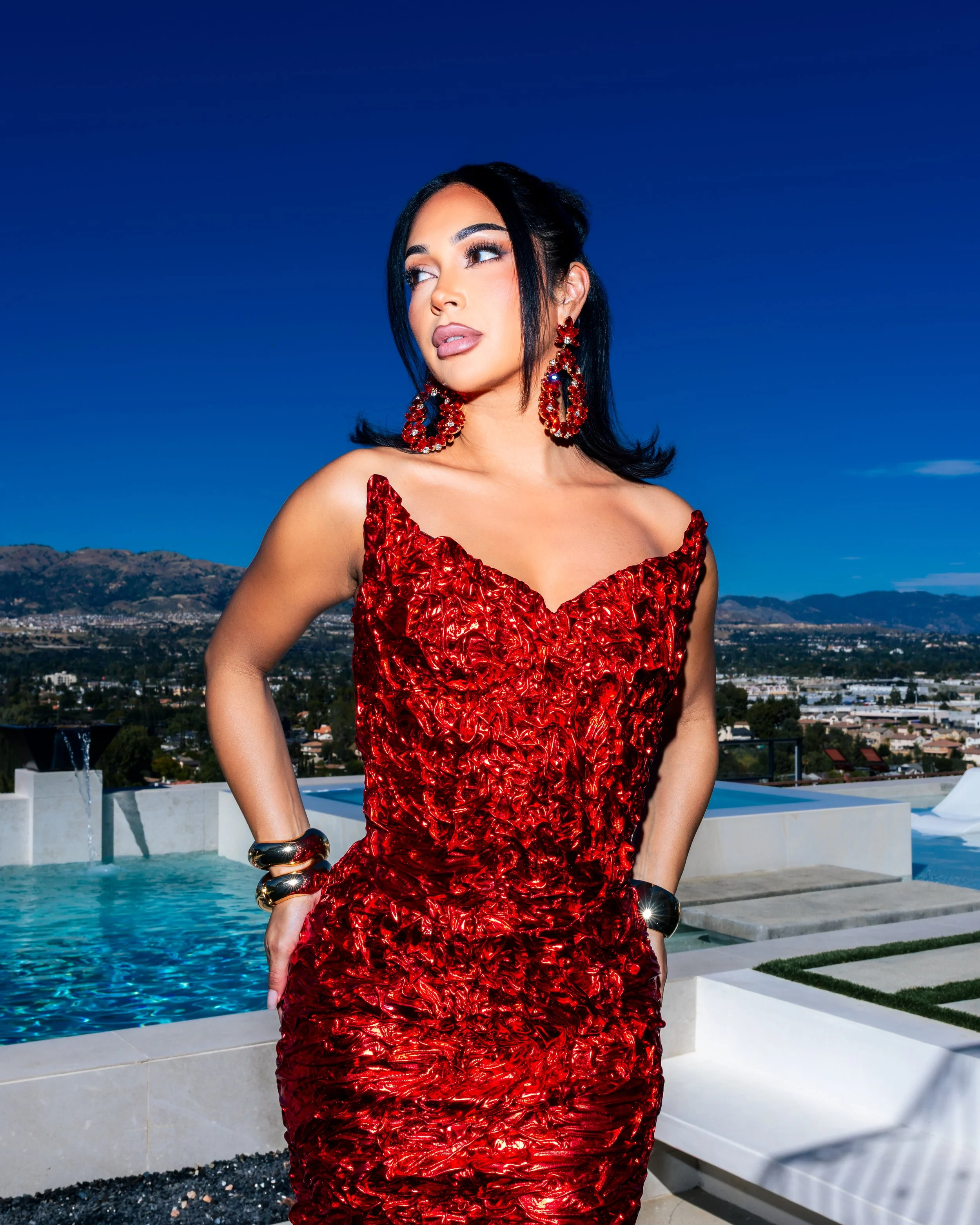 A woman in a red dress and matching earrings posing outdoors near a pool with a cityscape and mountains in the background.