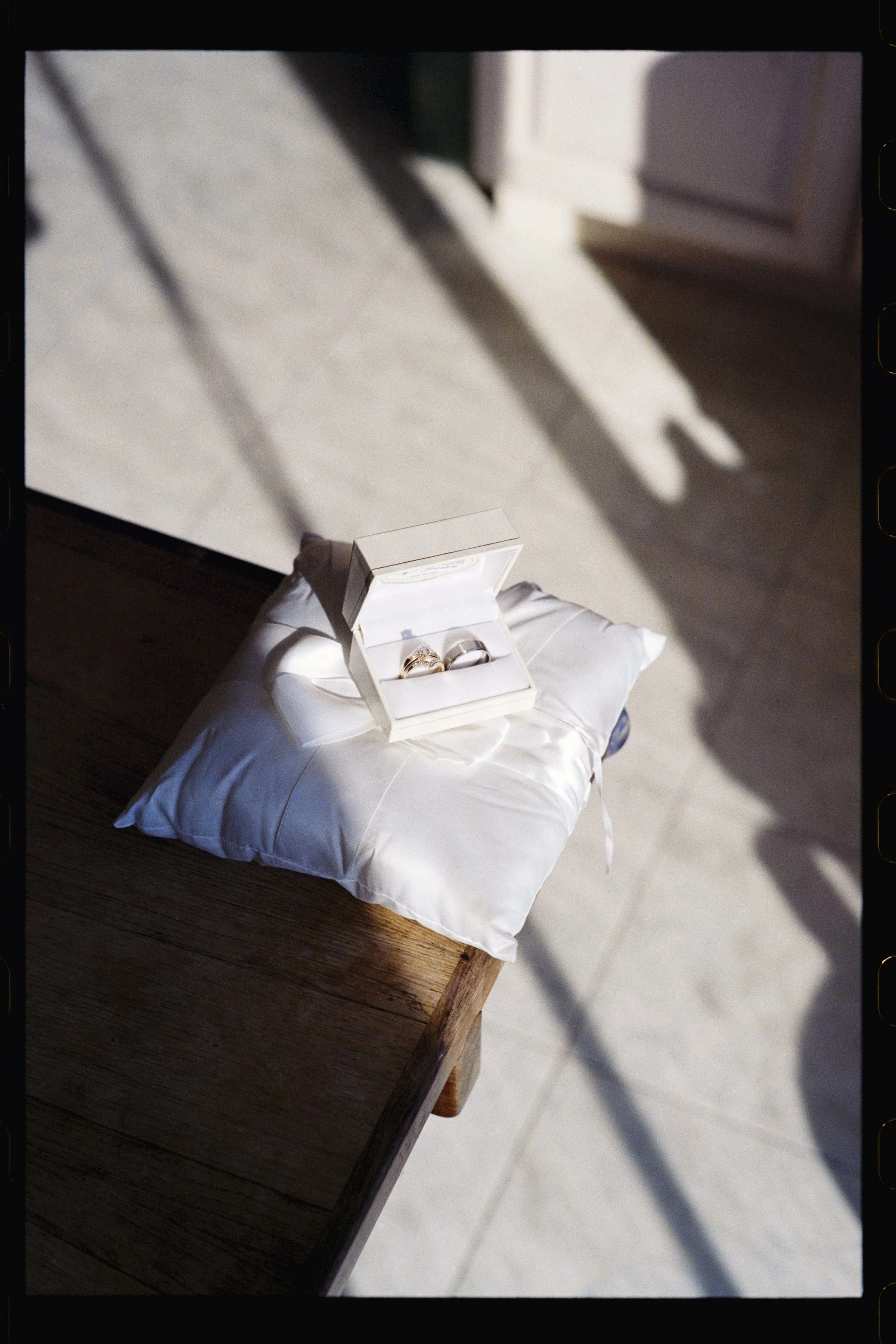 A white box with a lid, holding rings, is placed on a white pillow on a wooden table, with sunlight casting shadows.