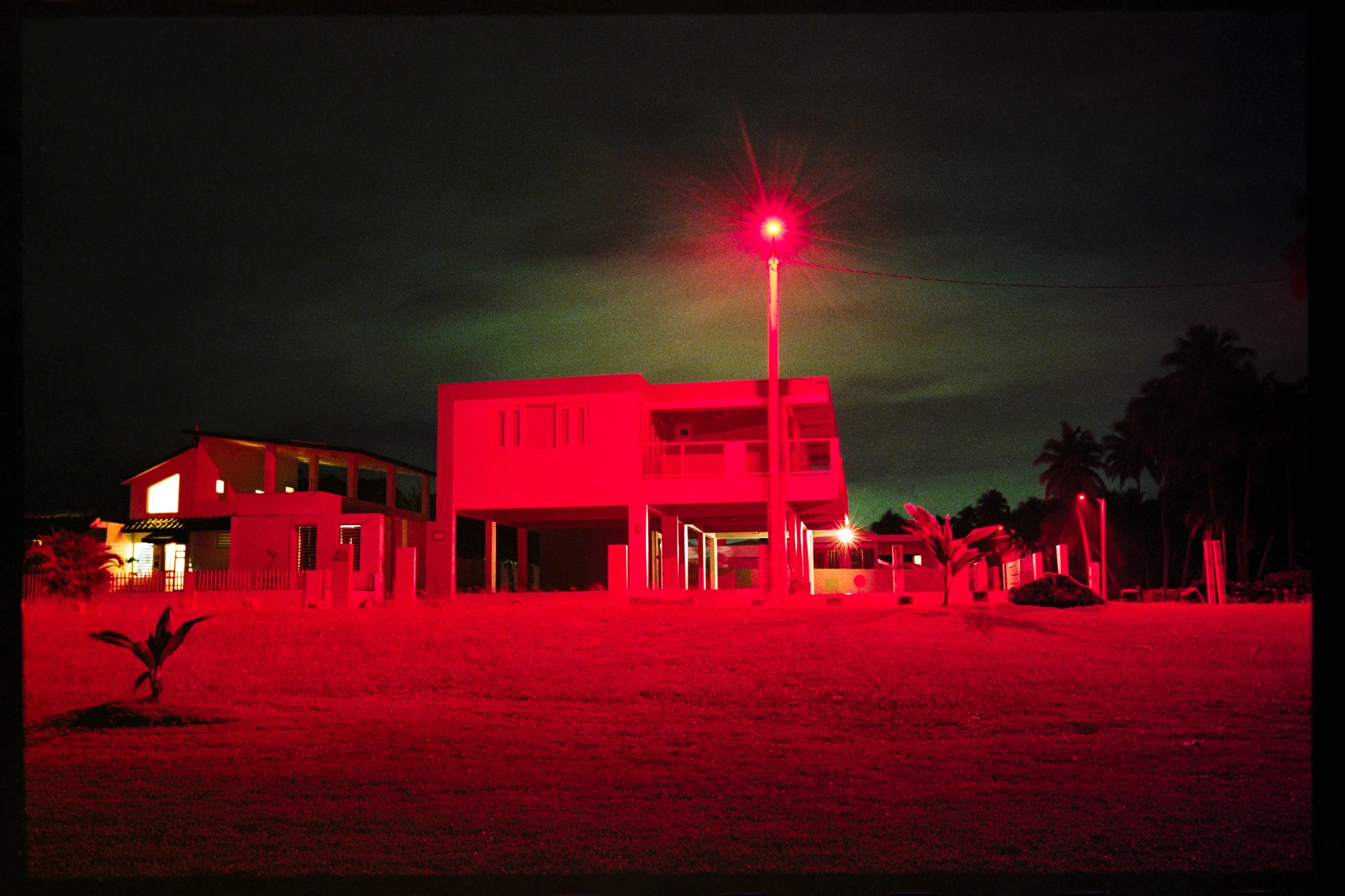 A nighttime scene of a modern building with a flat roof and open sides, illuminated by red light. A tall pole with a bright red light at the top is situated in front of the building. There are silhouettes of trees and plants nearby, and a dark sky wi