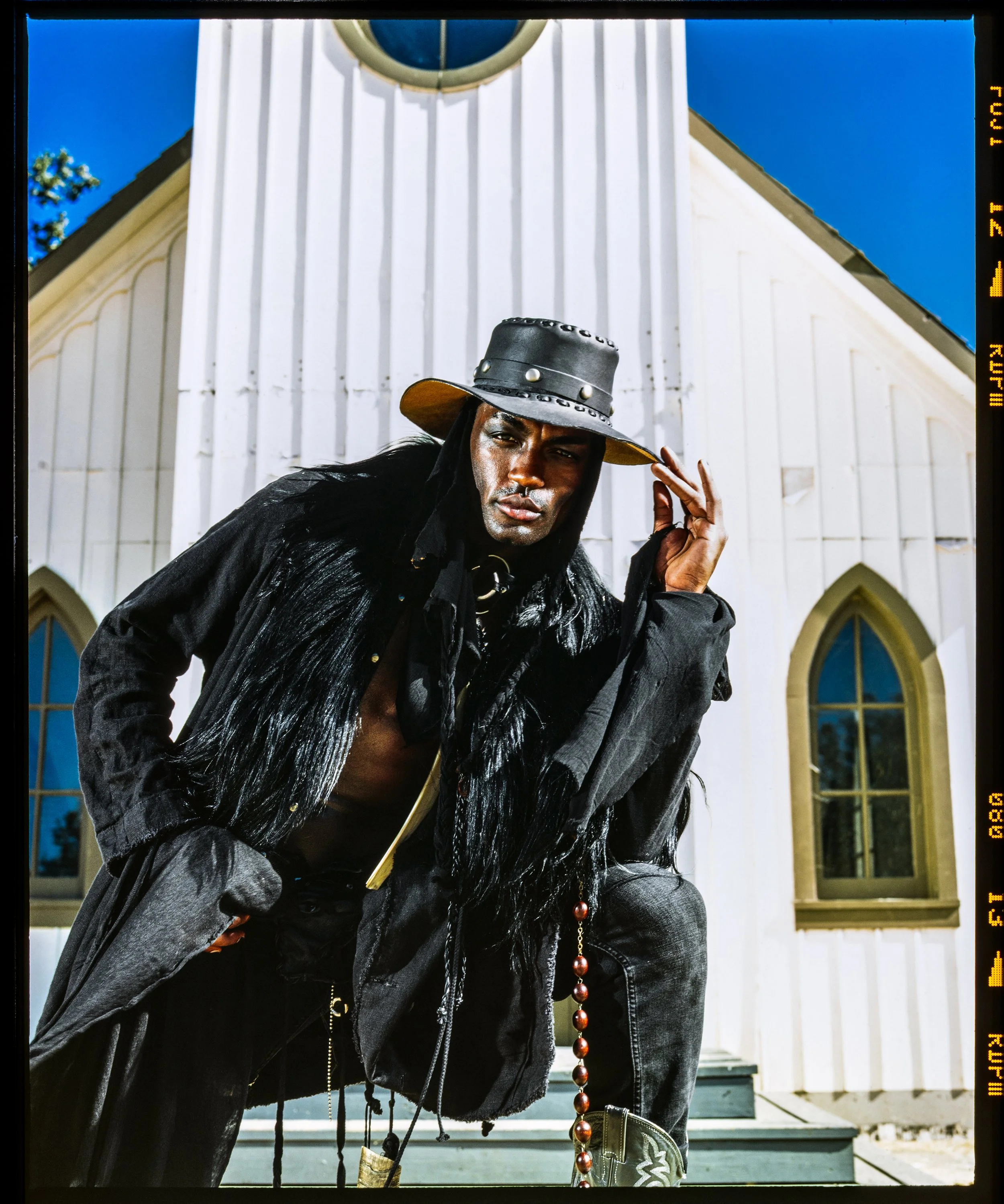 A man with dark hair and a serious expression, partly shirtless, wearing a black leather hat with metal details, and a long black coat with fur trim, standing outside in front of a white building with arched windows and a clock, under a clear blue sk