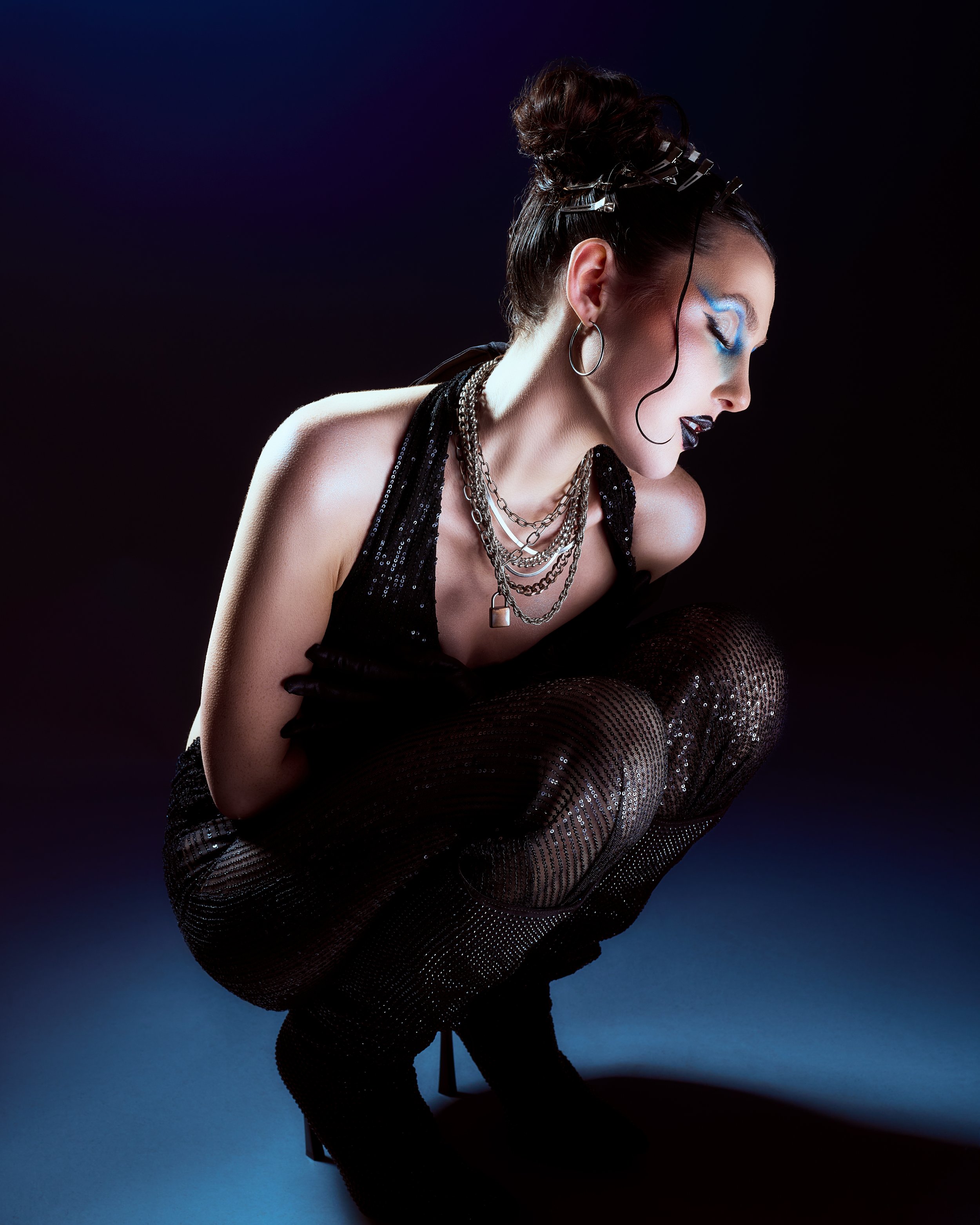 A woman crouching in a dark background with dramatic makeup and silver jewelry, wearing a black outfit and fishnet stockings.