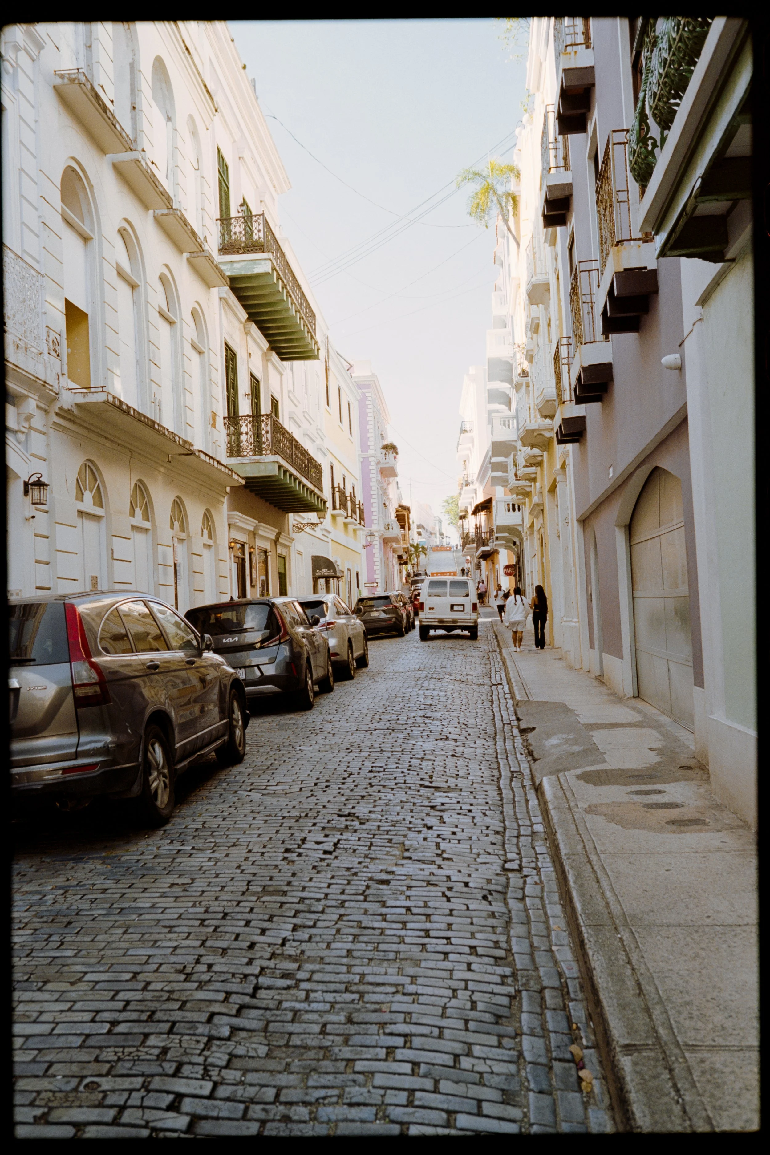 A narrow city street paved with cobblestones, lined with parked cars and tall, historic buildings with balconies and large windows. Pedestrians walk along the sidewalk on the right side of the street. The scene is illuminated by daylight with a clear
