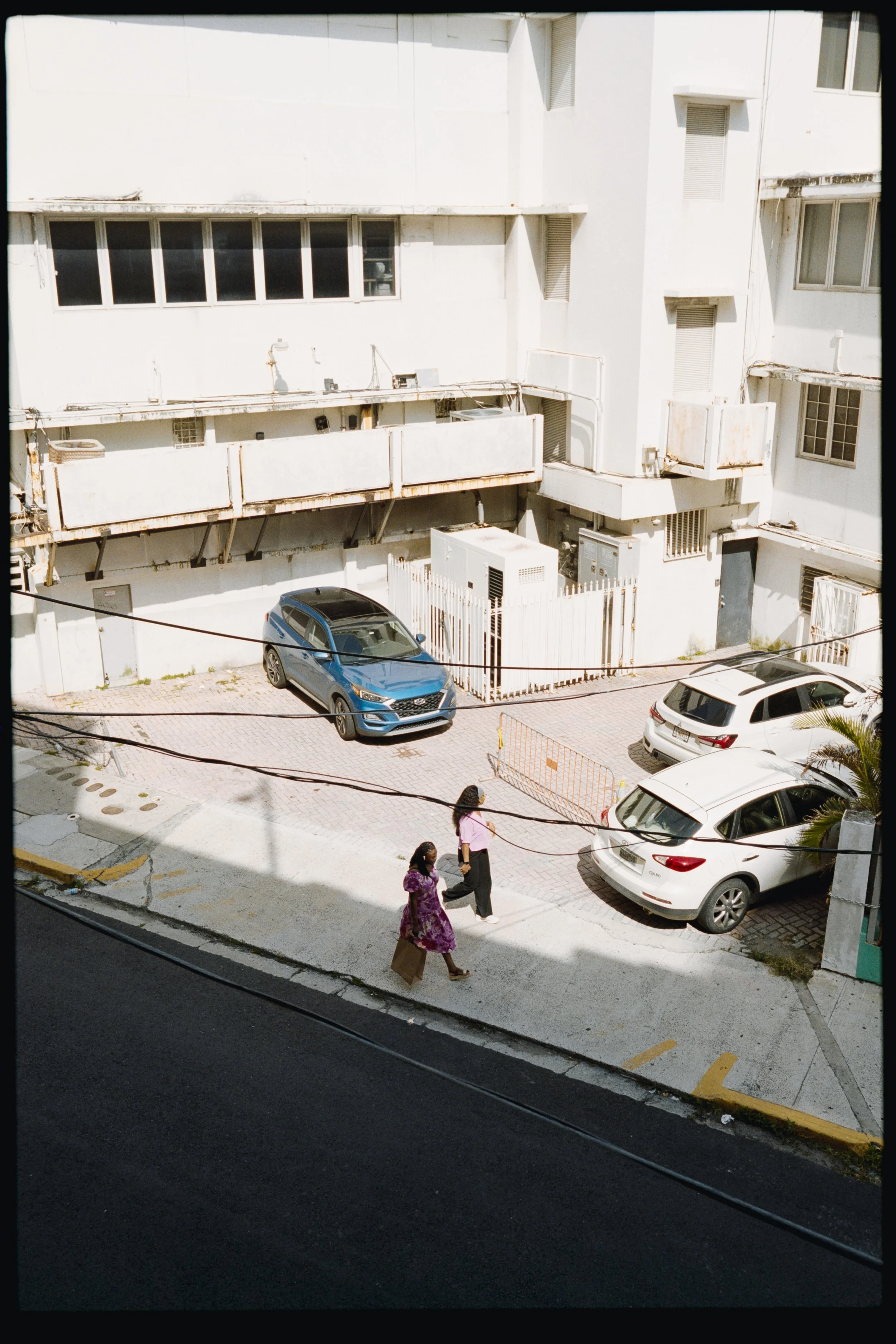 A sunny urban street scene with three women walking along the sidewalk, two dressed in traditional pink and purple dresses, and one in black pants and a grey shirt, carrying shopping bags. There are several parked cars including a blue SUV and a whit