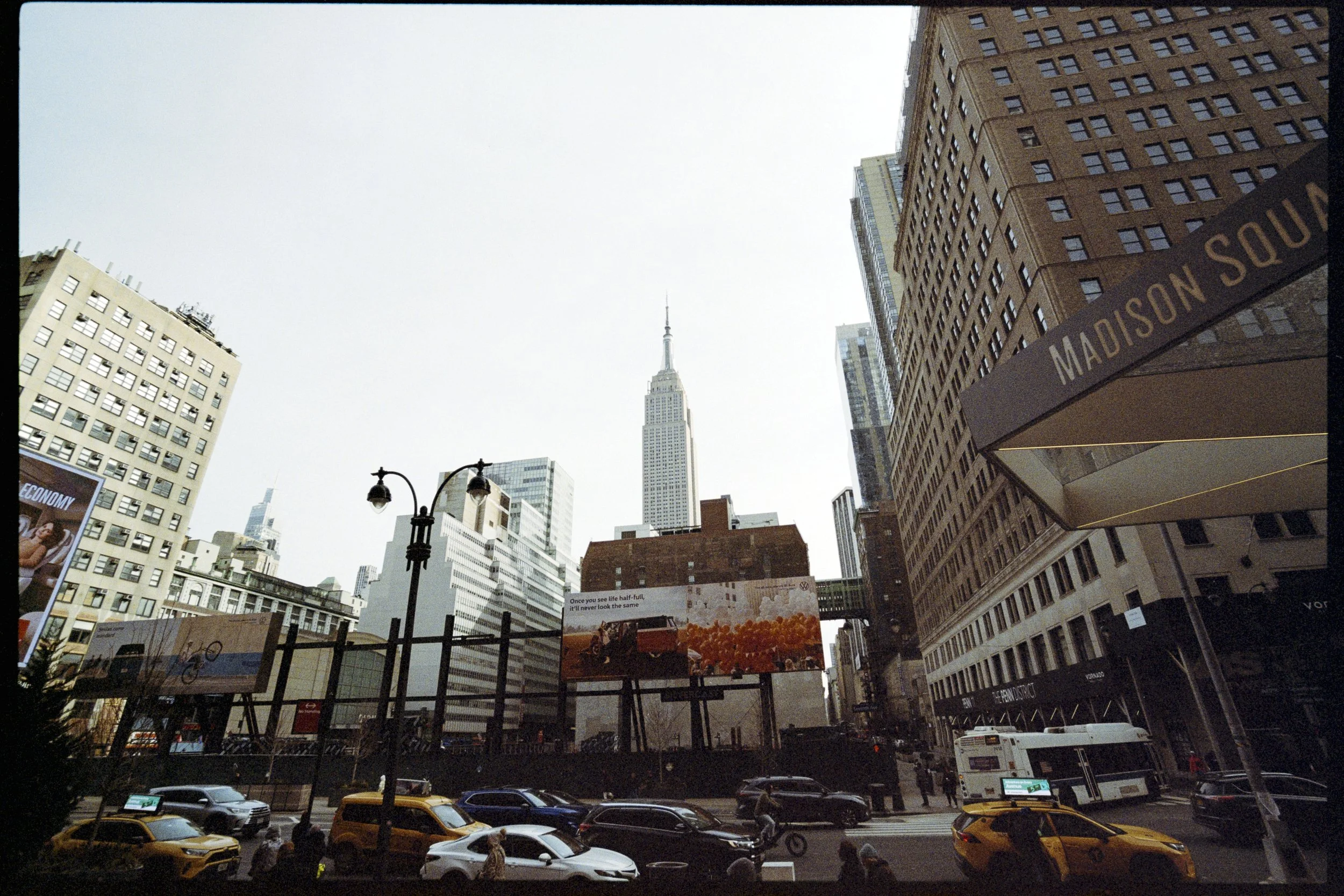 A busy city street in New York City with tall buildings, a billboard, cars parked on the street, and the Empire State Building visible in the background under a cloudy sky.