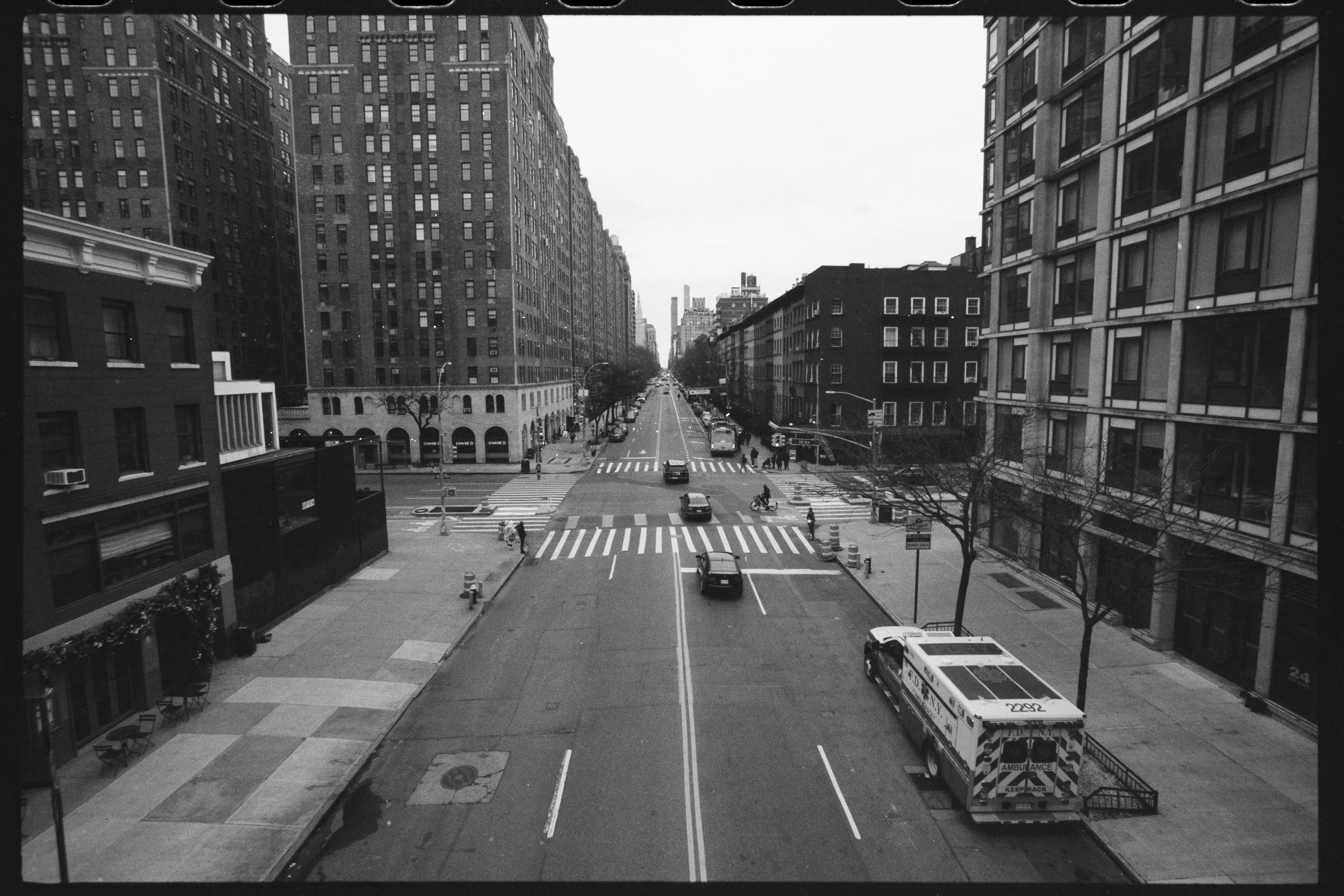 Black and white photo of an urban city street with tall buildings on both sides. The street has a crosswalk, a few cars, people walking, and an ambulance parked near the sidewalk.