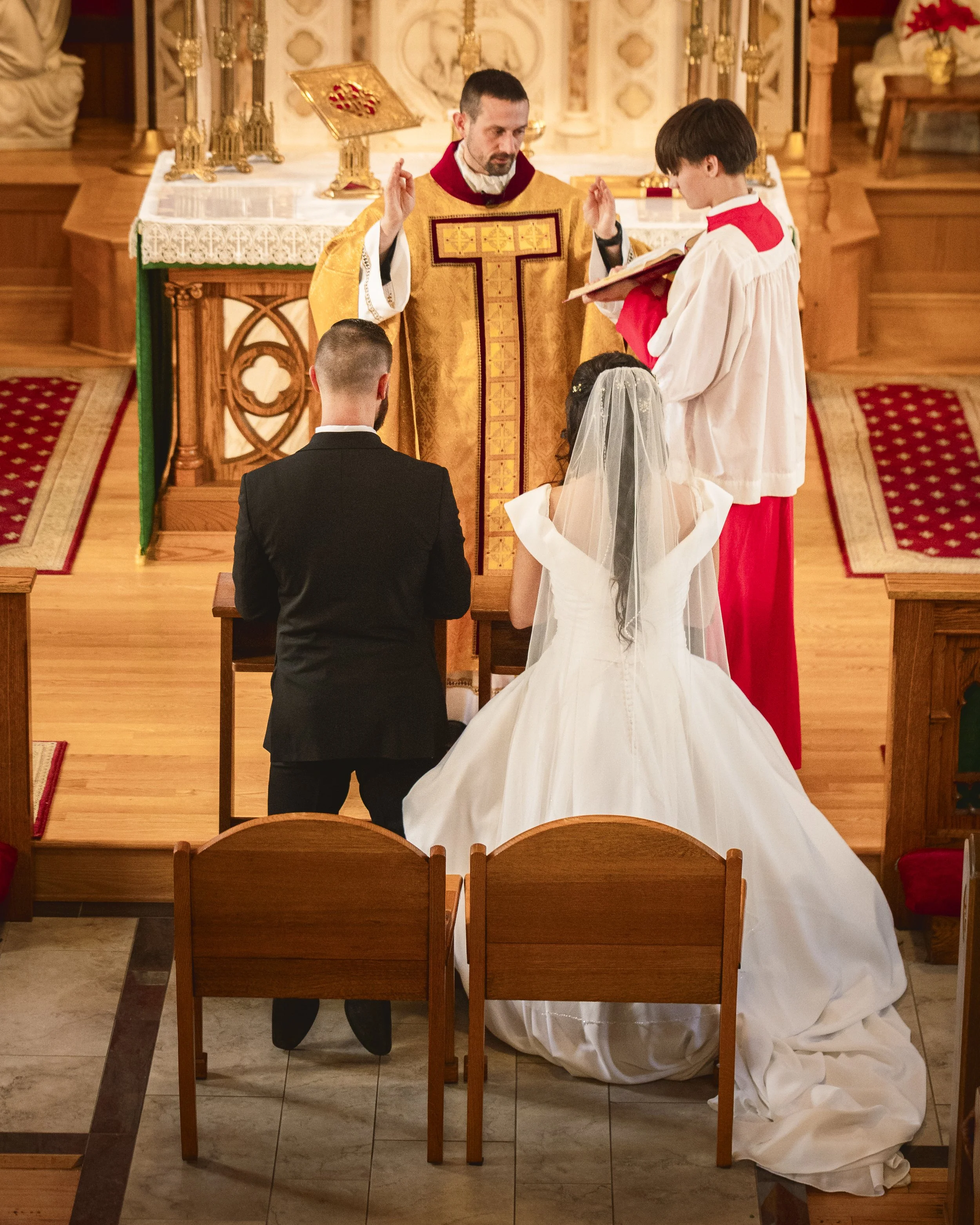 A priest officiates a wedding ceremony in a church. The bride, in a white wedding gown with a veil, and the groom, in a black suit, kneel before the altar. An altar server, in a white robe with red accents, holds a book to read from. The altar is dec