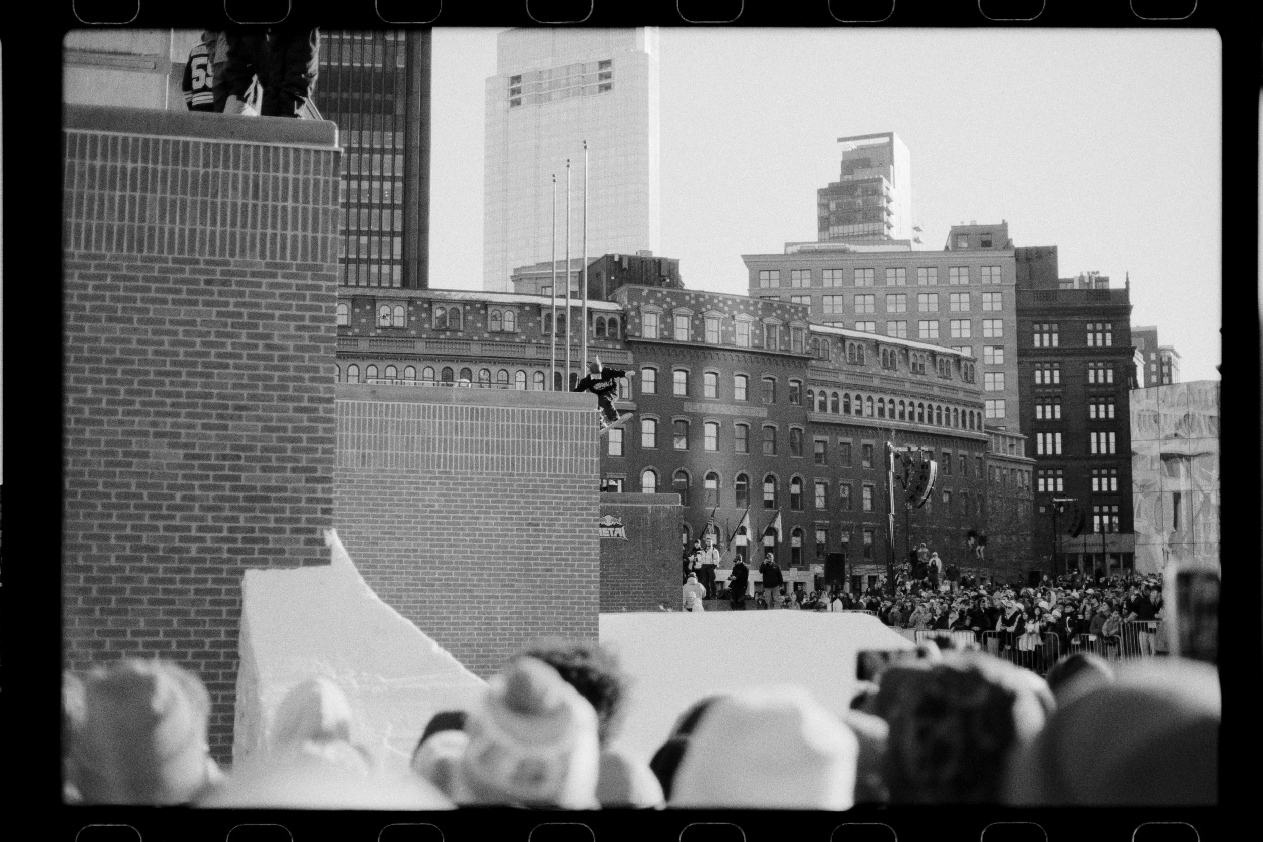 Black and white photo of a ski jump with a wooden ramp in the foreground and a cityscape with tall buildings and a crowd below in the background.