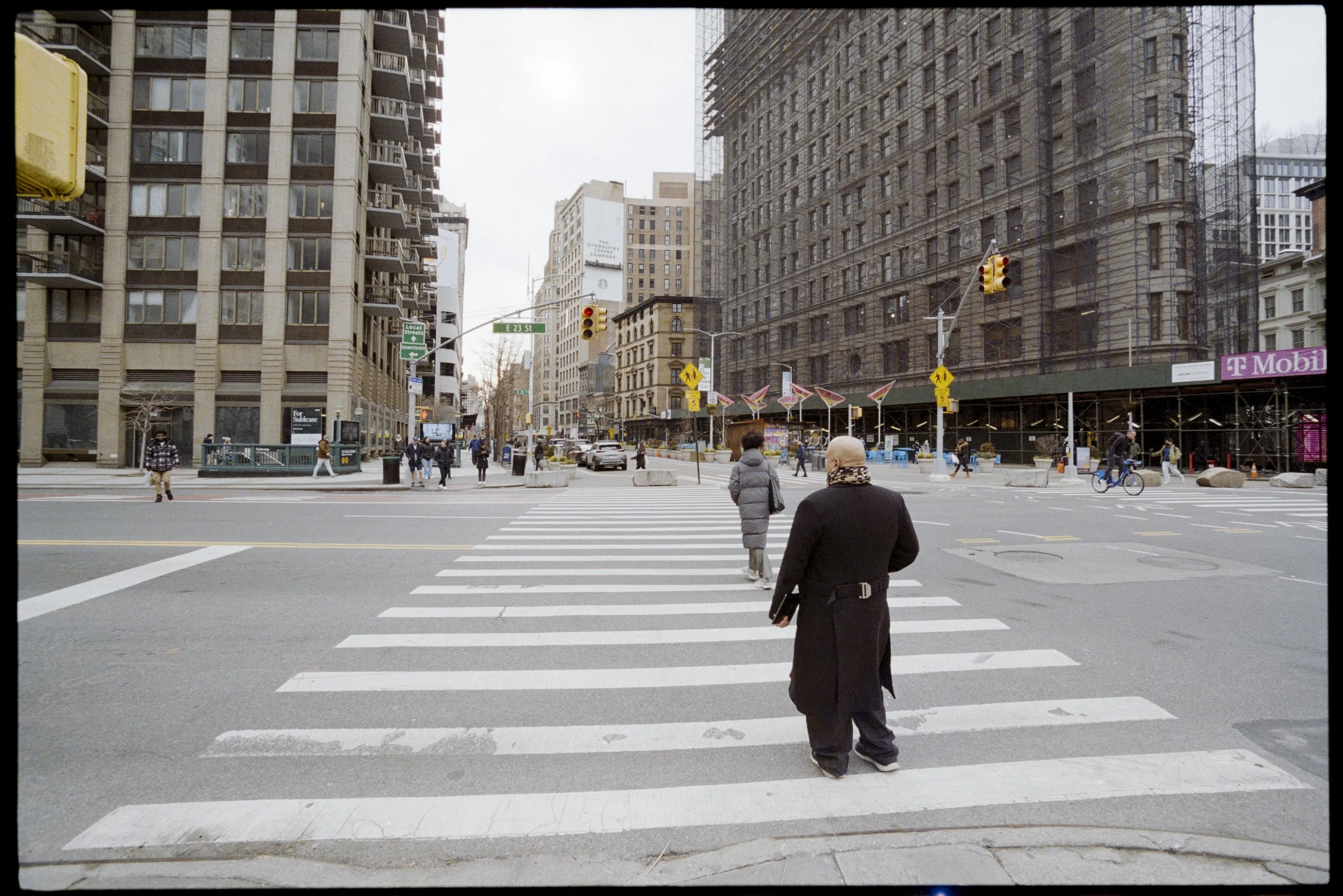 City street scene with pedestrians crossing at a crosswalk, tall buildings, and traffic lights, on a cloudy day.