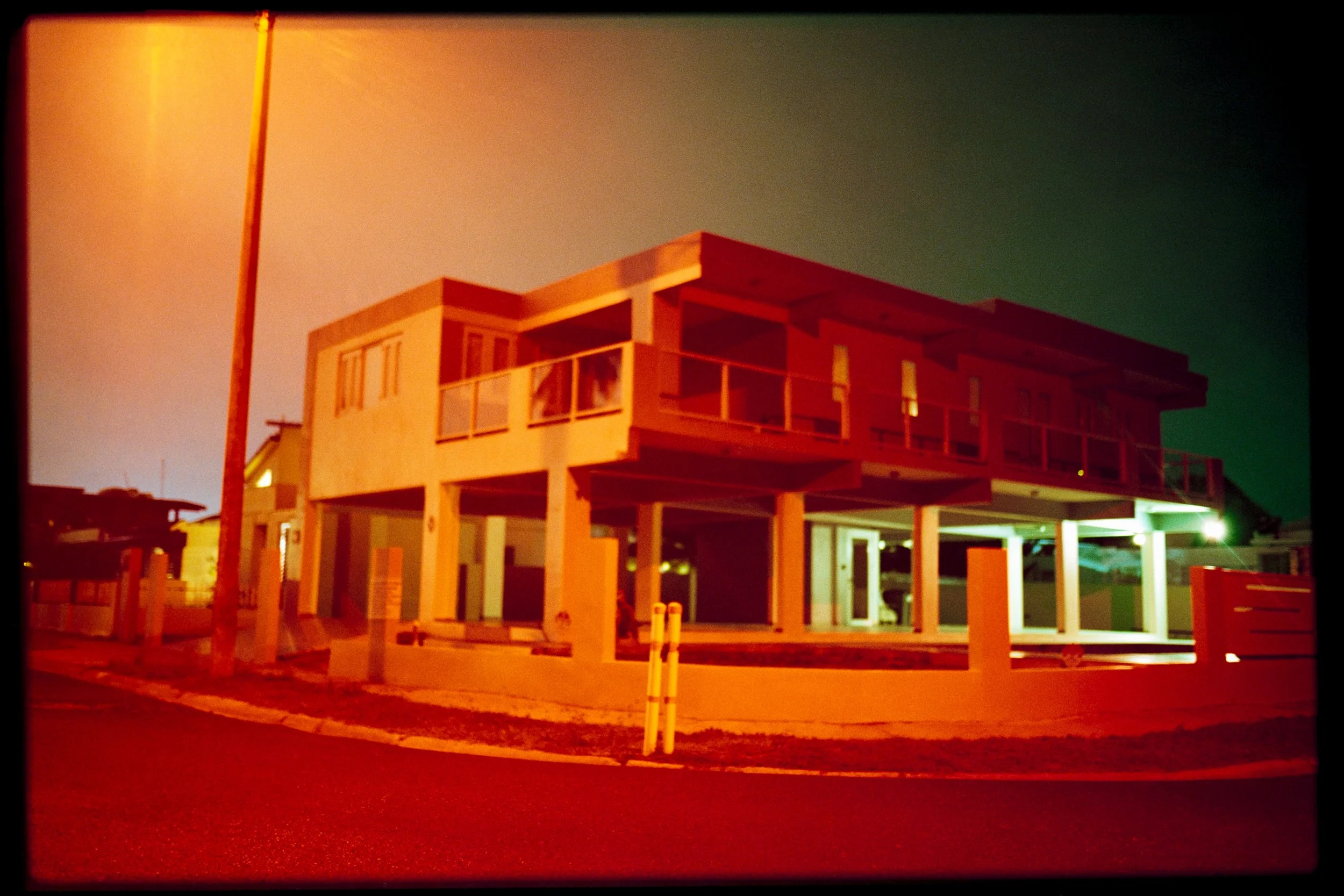 A modern, multi-story house under construction with exposed wooden framing, situated at night with colored lights illuminating the structure.