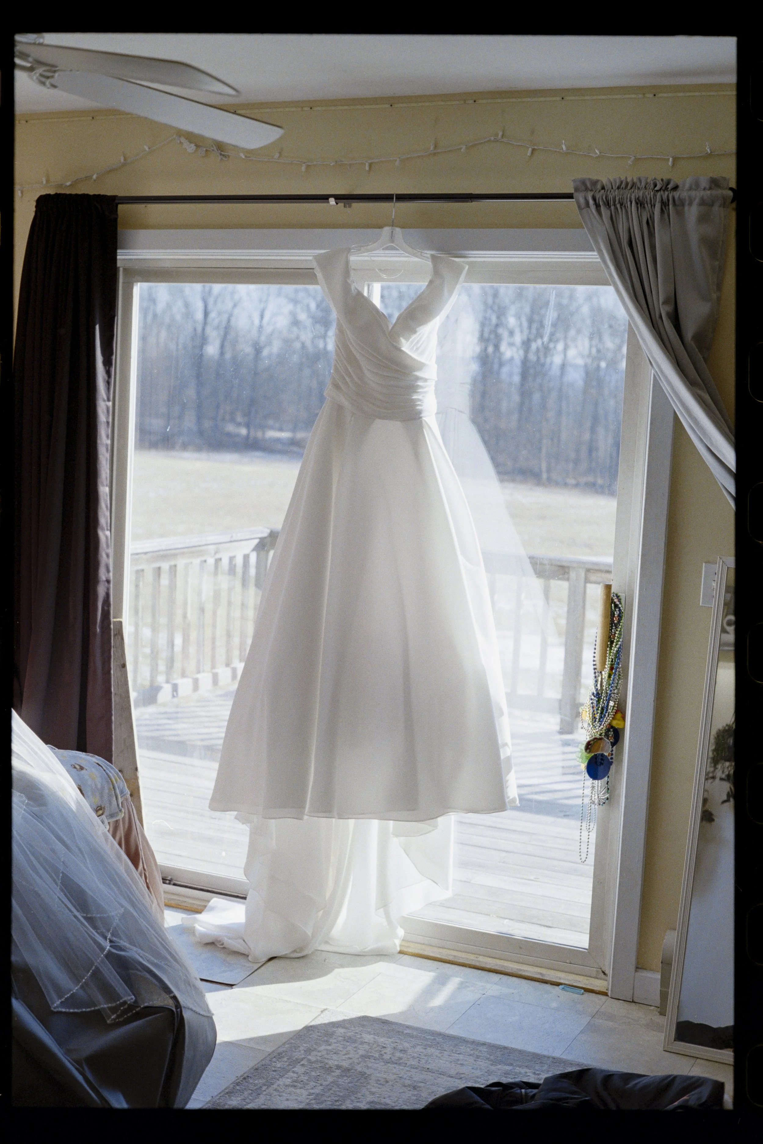 A white wedding dress hanging on a clear plastic hanger inside a room by a sliding glass door with gray curtains, outside view of trees and cloudy sky.