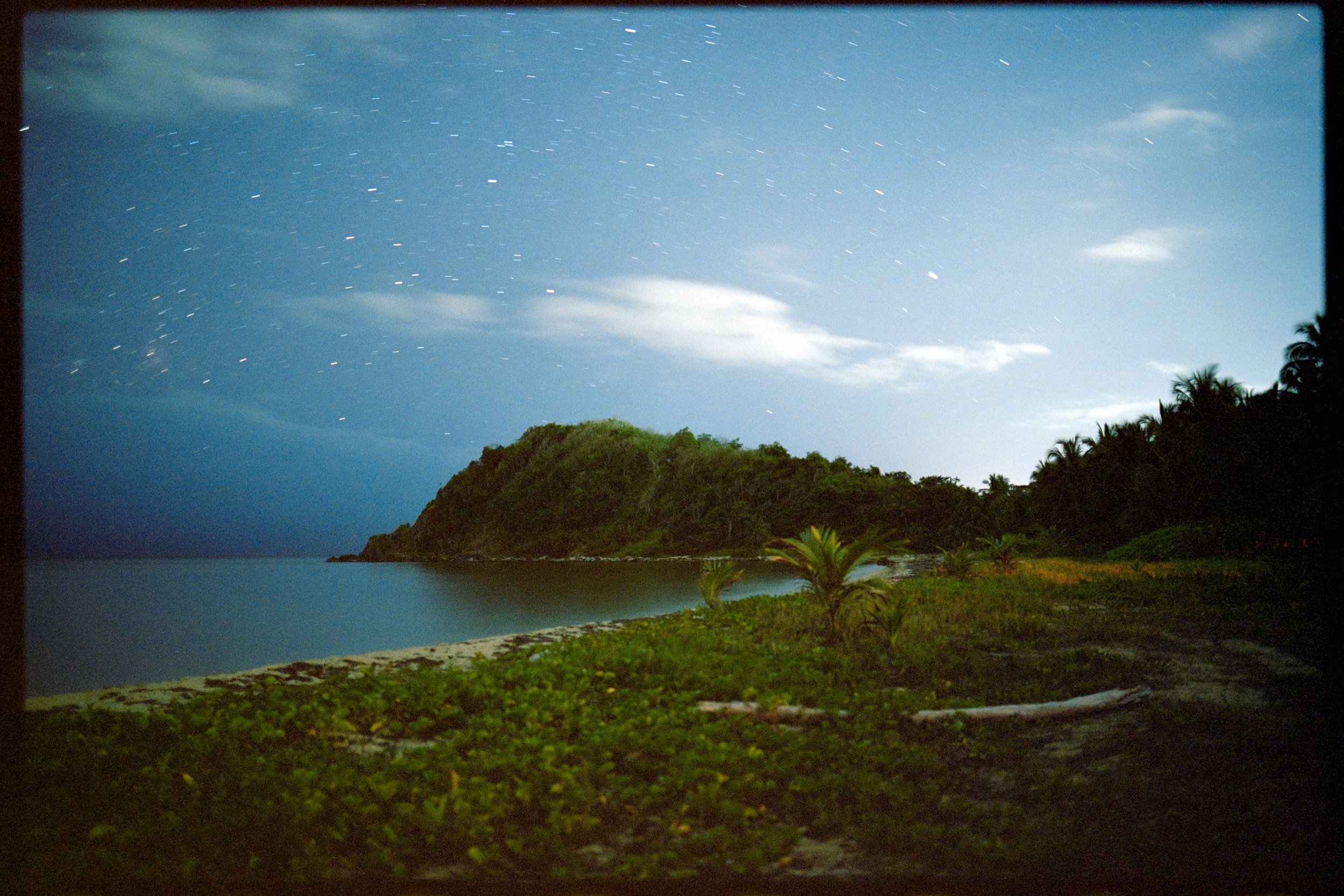 Nighttime view of a tropical coastline with lush green hills, palm trees, a small beach, calm water, and a starry sky with some clouds.