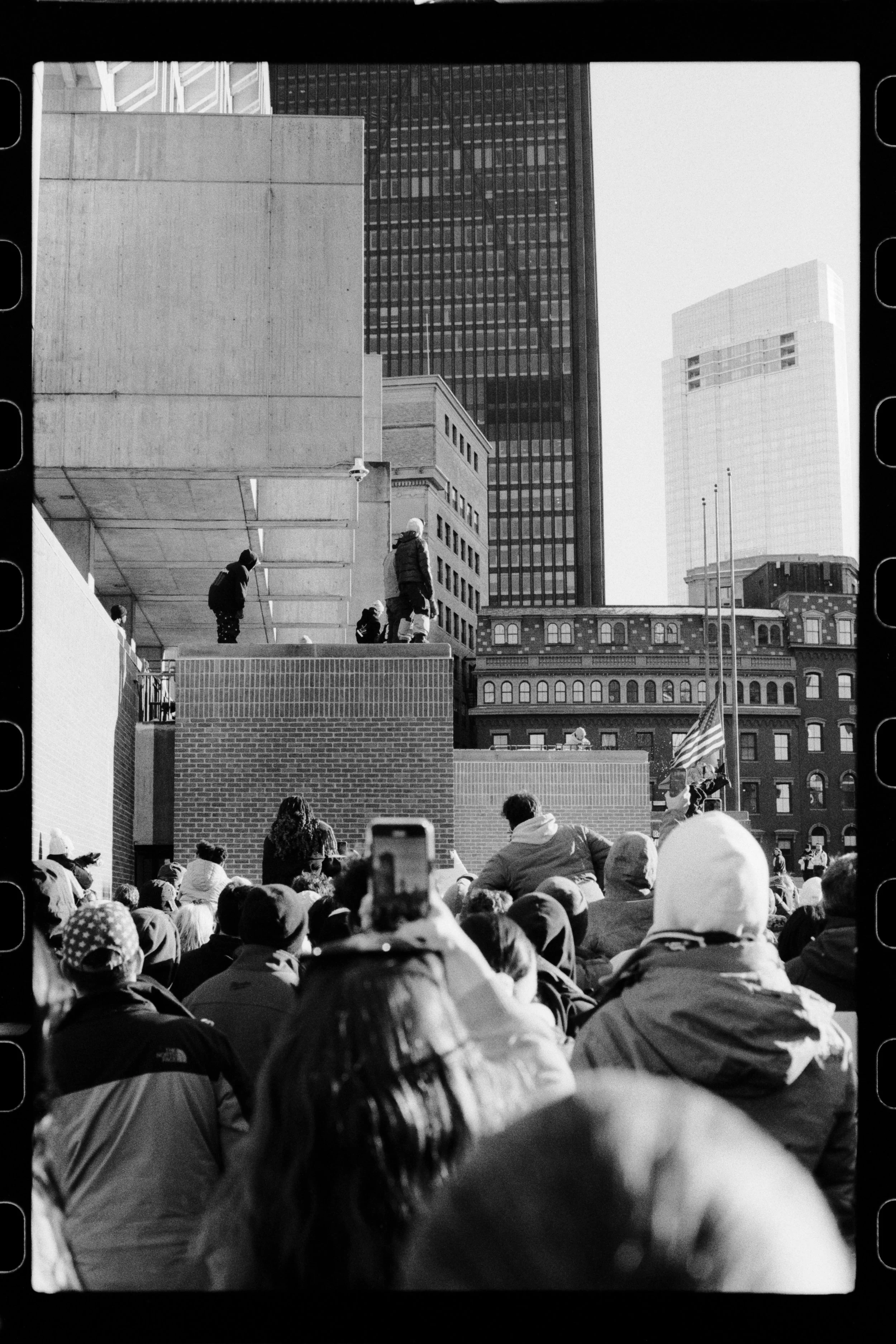 A crowd of people gathered outdoors on a sunny day, facing a stage or platform with several individuals standing and speaking or performing in front of a backdrop of tall city buildings.