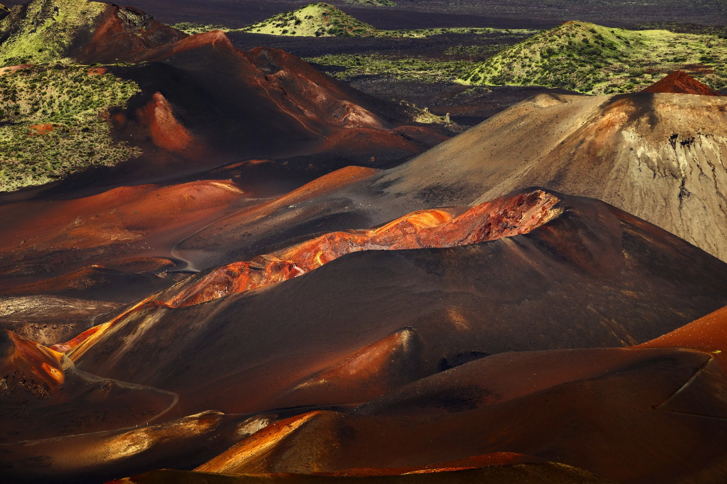 Colorful volcanic landscape with various hills and mounds in shades of red, brown, gray, and green, with some green vegetation on the hills and dark volcanic soil.