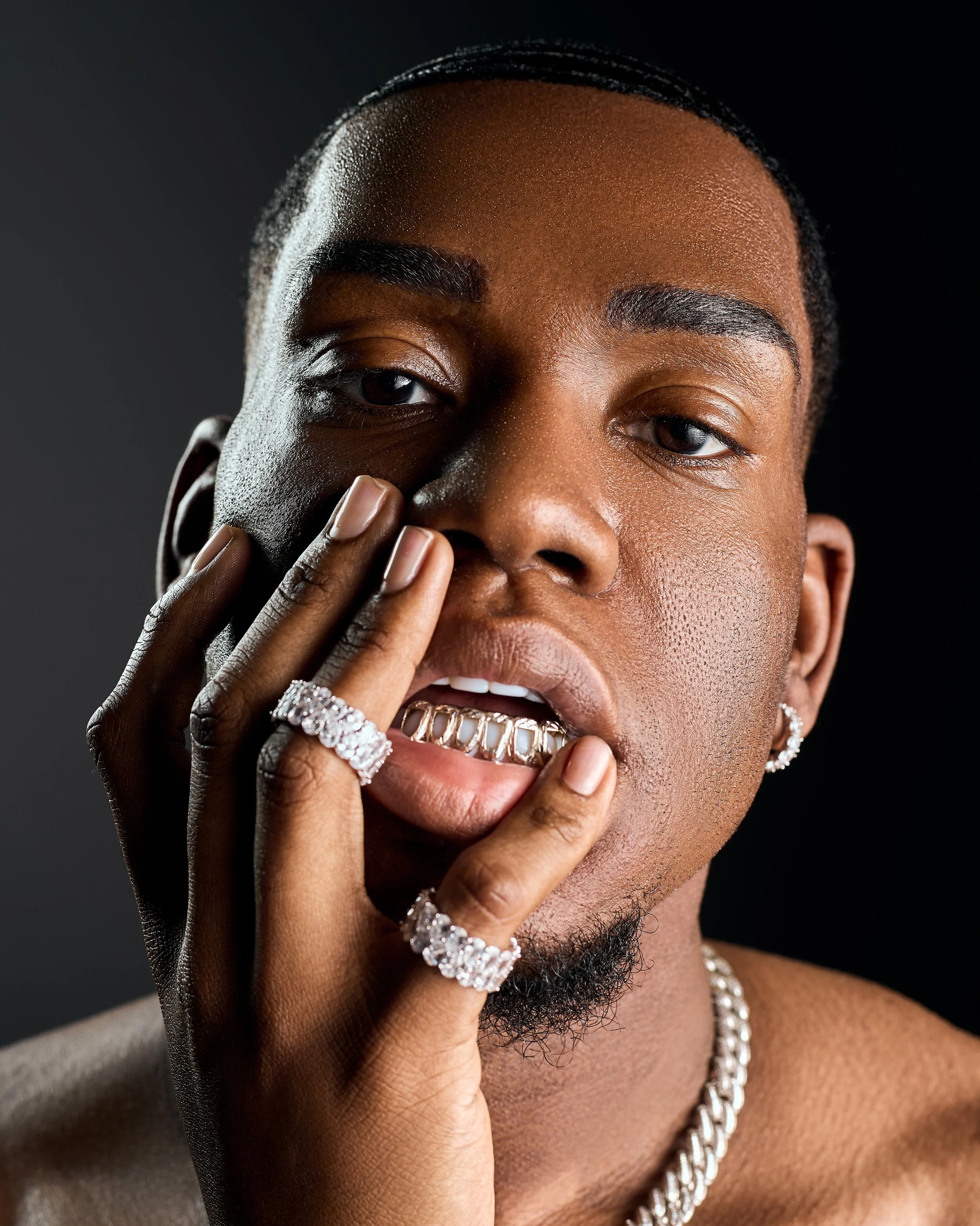 Close-up portrait of a Black man with jewelry, touching his face with his hand, against a dark background.
