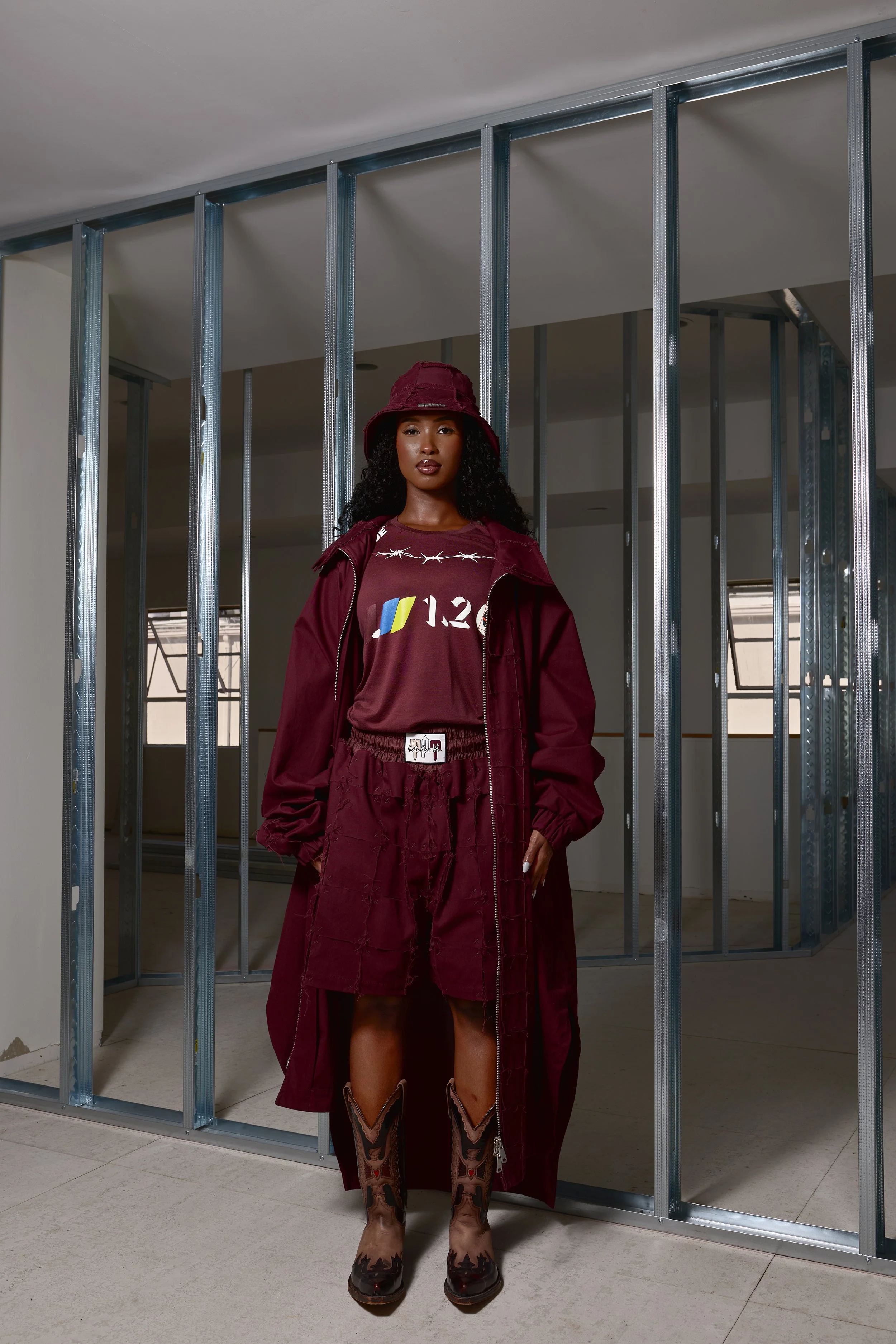 A woman dressed in a burgundy outfit, including a shirt, jacket, hat, and skirt, standing in front of a construction frame inside a building.