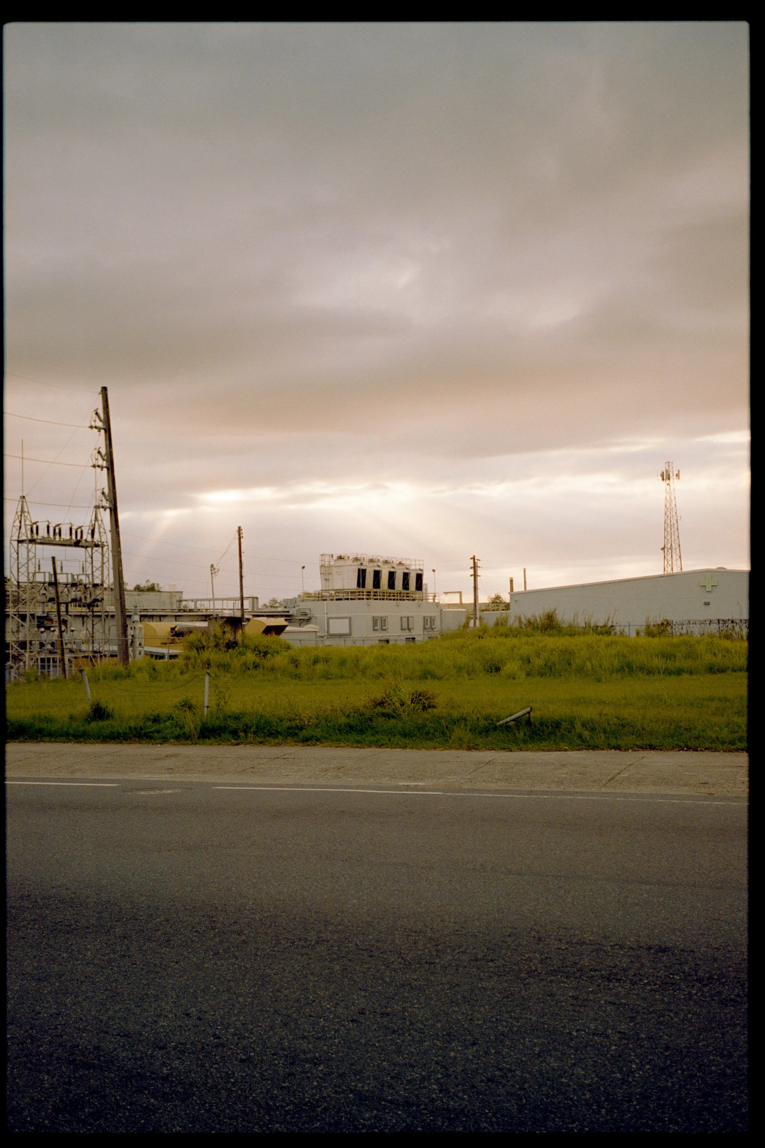 View of a cloudy sky over an industrial area with multiple buildings, power lines, and a grassy field in the foreground. The scene is captured during dawn or dusk.
