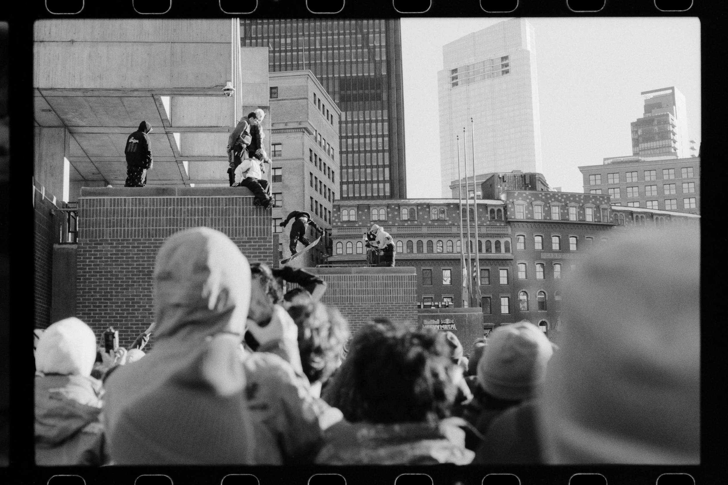 Black and white photo of a crowd at an outdoor event, looking at a stage with people climbing on a wall structure in an urban setting with tall buildings in the background.