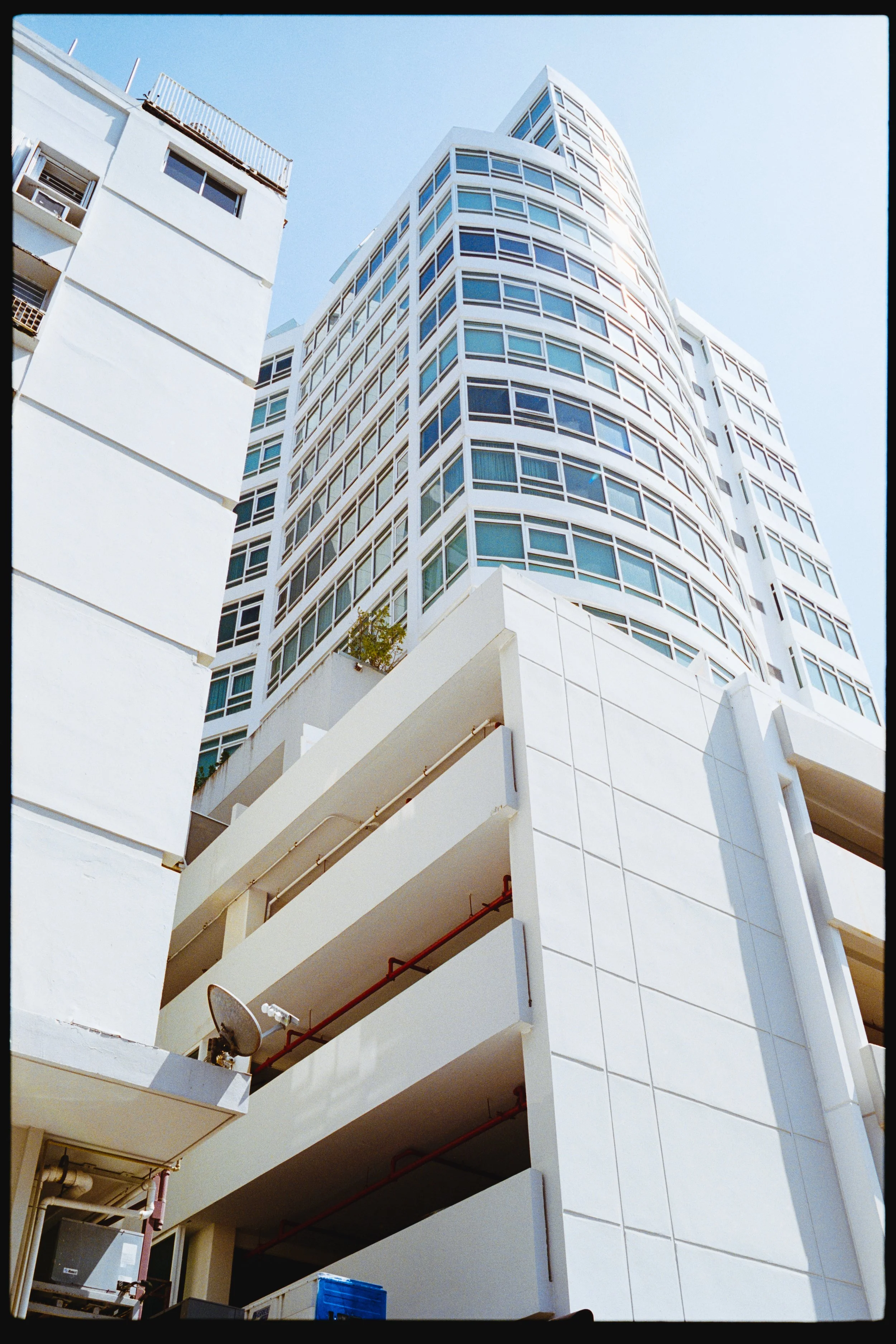White modern high-rise building with multiple windows and a curved corner, viewed from below against a clear blue sky.