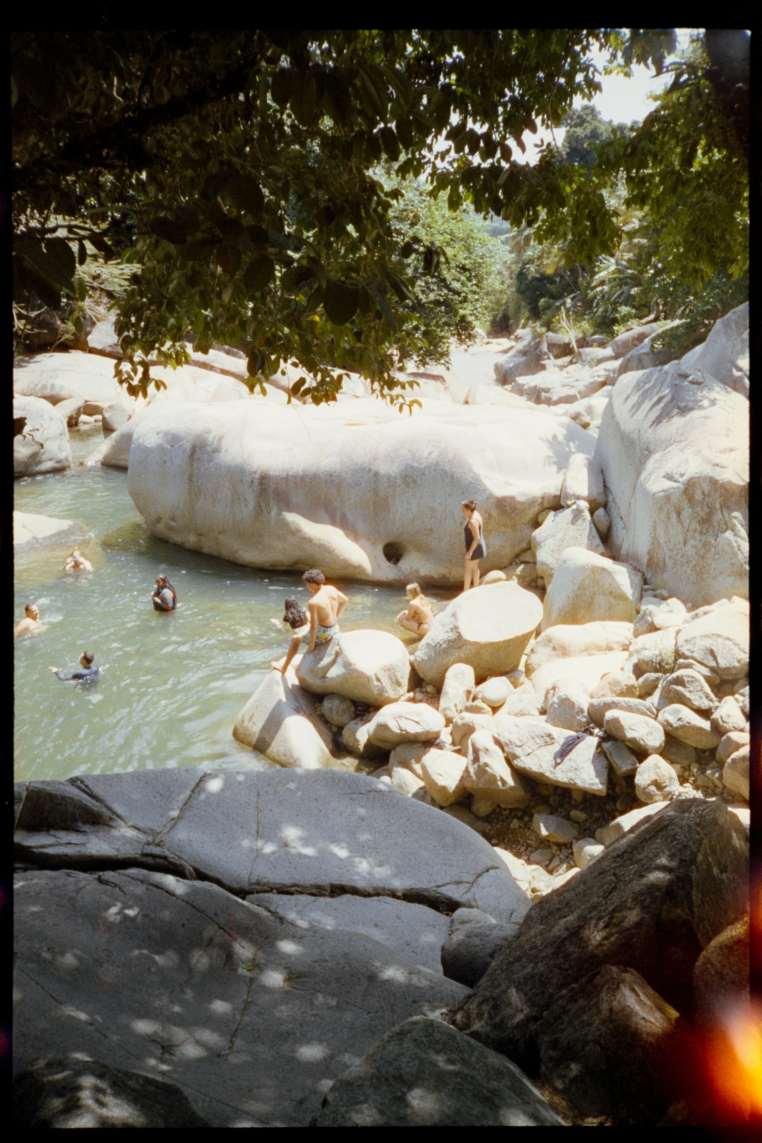 People swimming and sitting on rocks in a river surrounded by trees and large boulders.