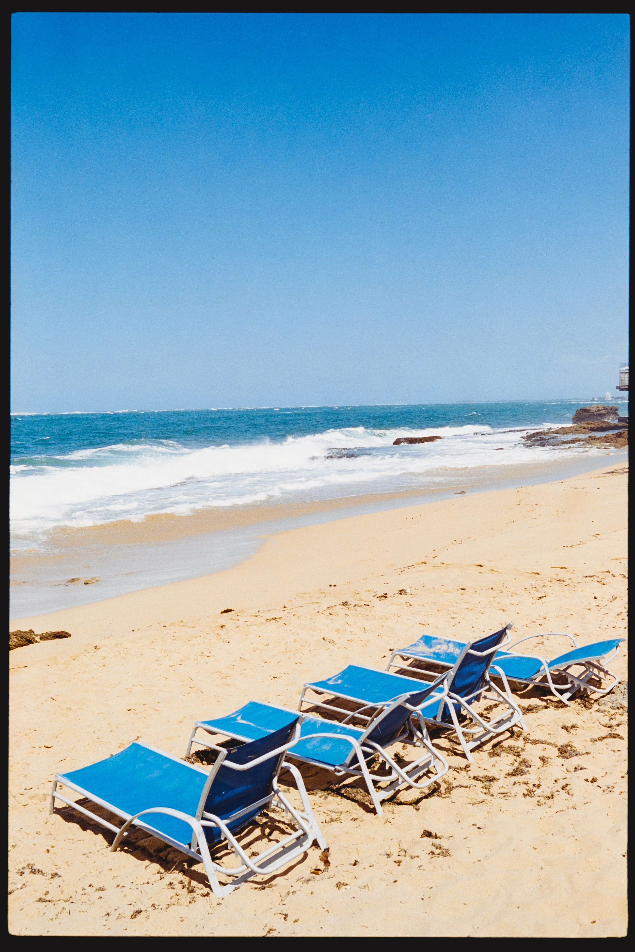 Beach with four blue lounge chairs on sand, ocean waves, rocks on the shoreline, and buildings in the background under a clear blue sky.