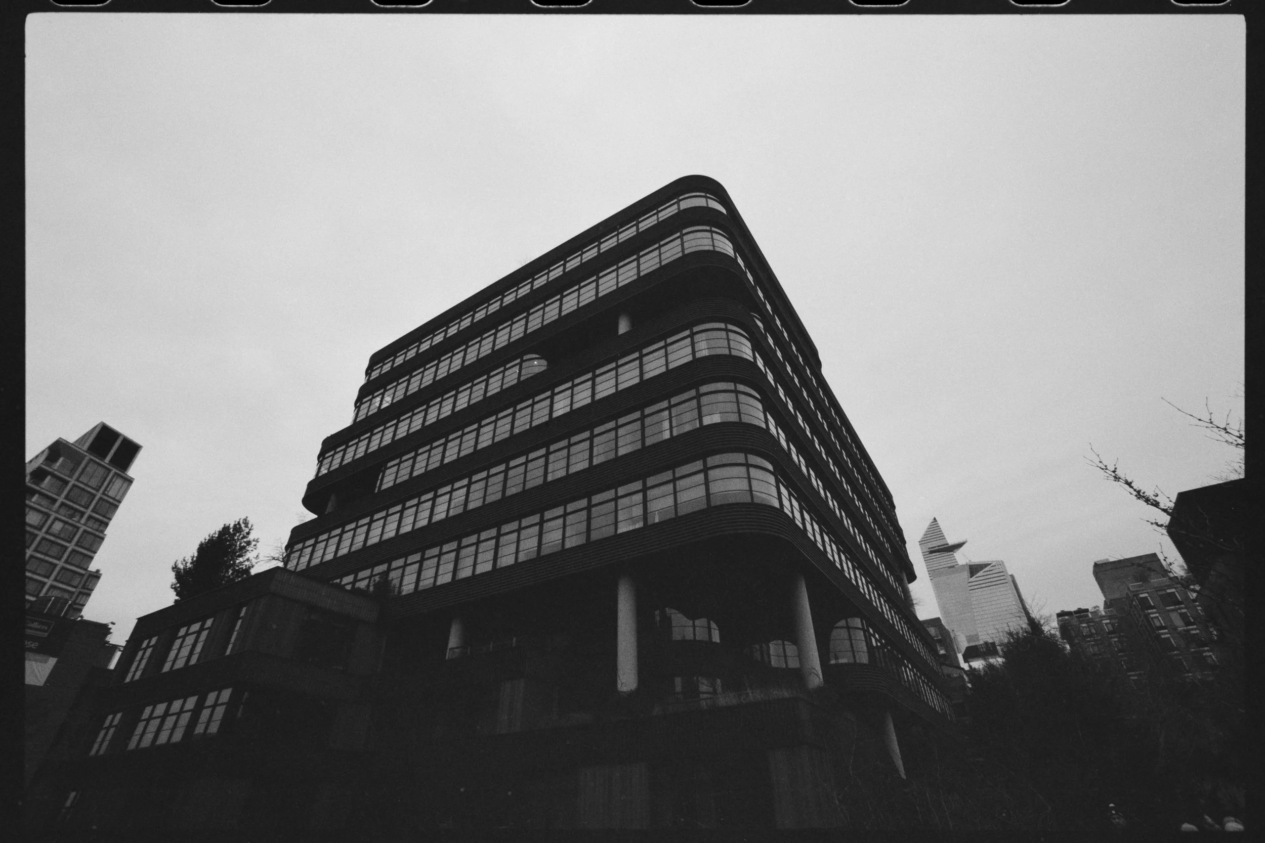 Black and white photo of a modern multi-story building with rounded edges and glass windows, surrounded by other high-rise buildings, with some trees and a cloudy sky in the background.