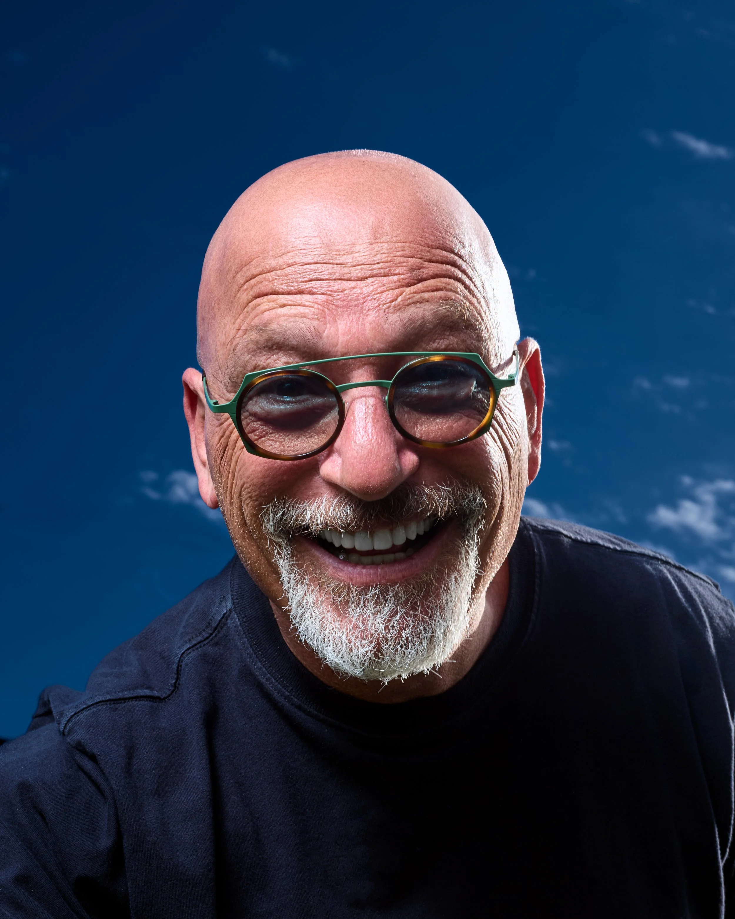 A smiling, bald man with a white beard wearing glasses and a black shirt, against a clear blue sky with some clouds.