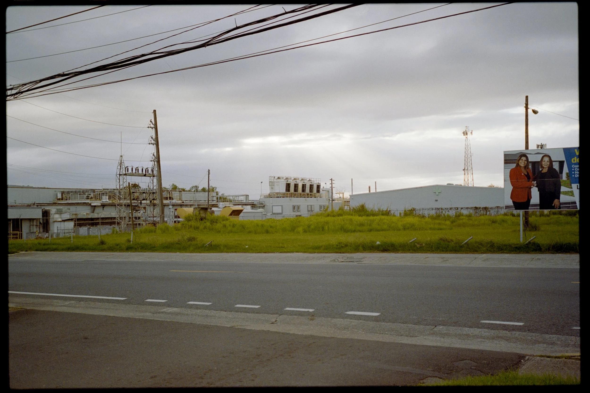 View of industrial buildings and power lines under a cloudy sky, with a billboard featuring two women shaking hands on the right side.
