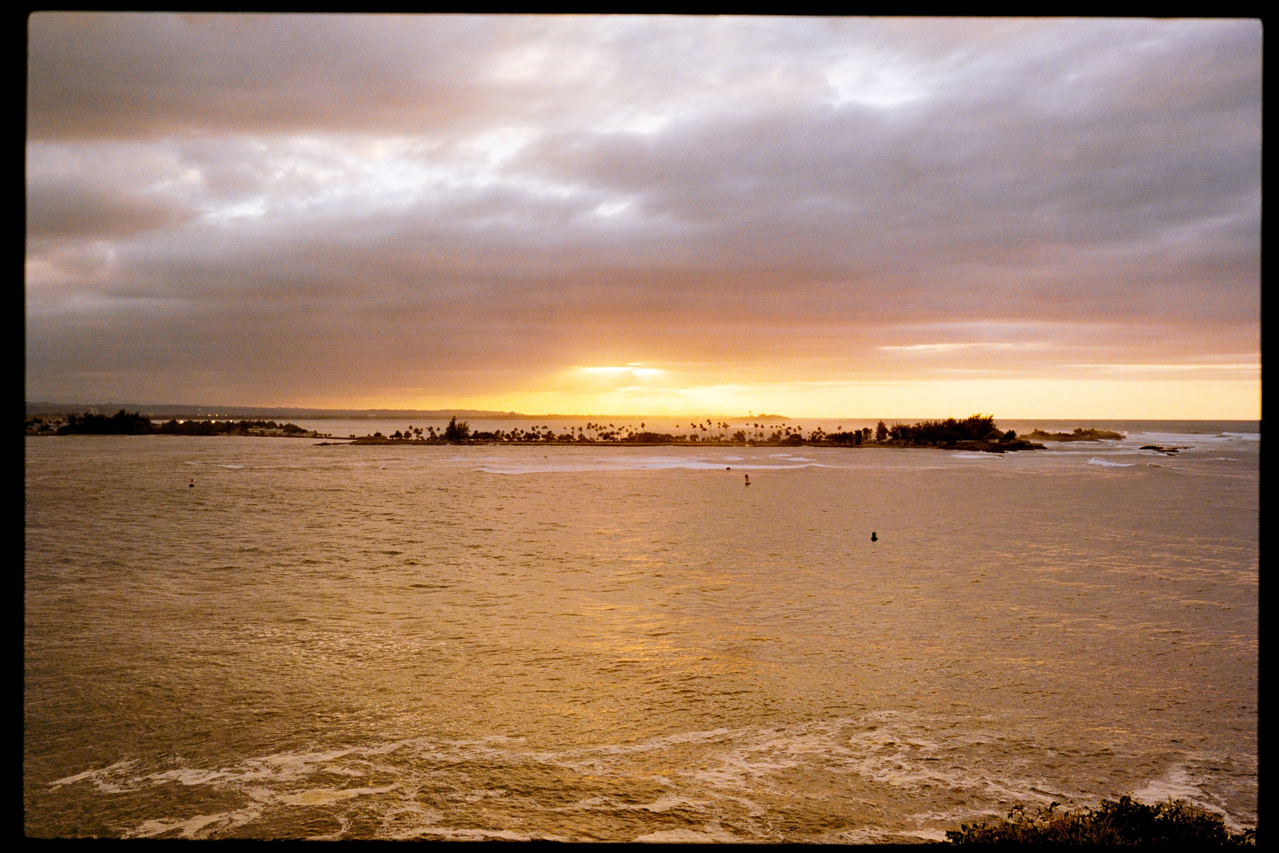 Sunset over a calm body of water with a cloudy sky and distant shoreline lined with palm trees.