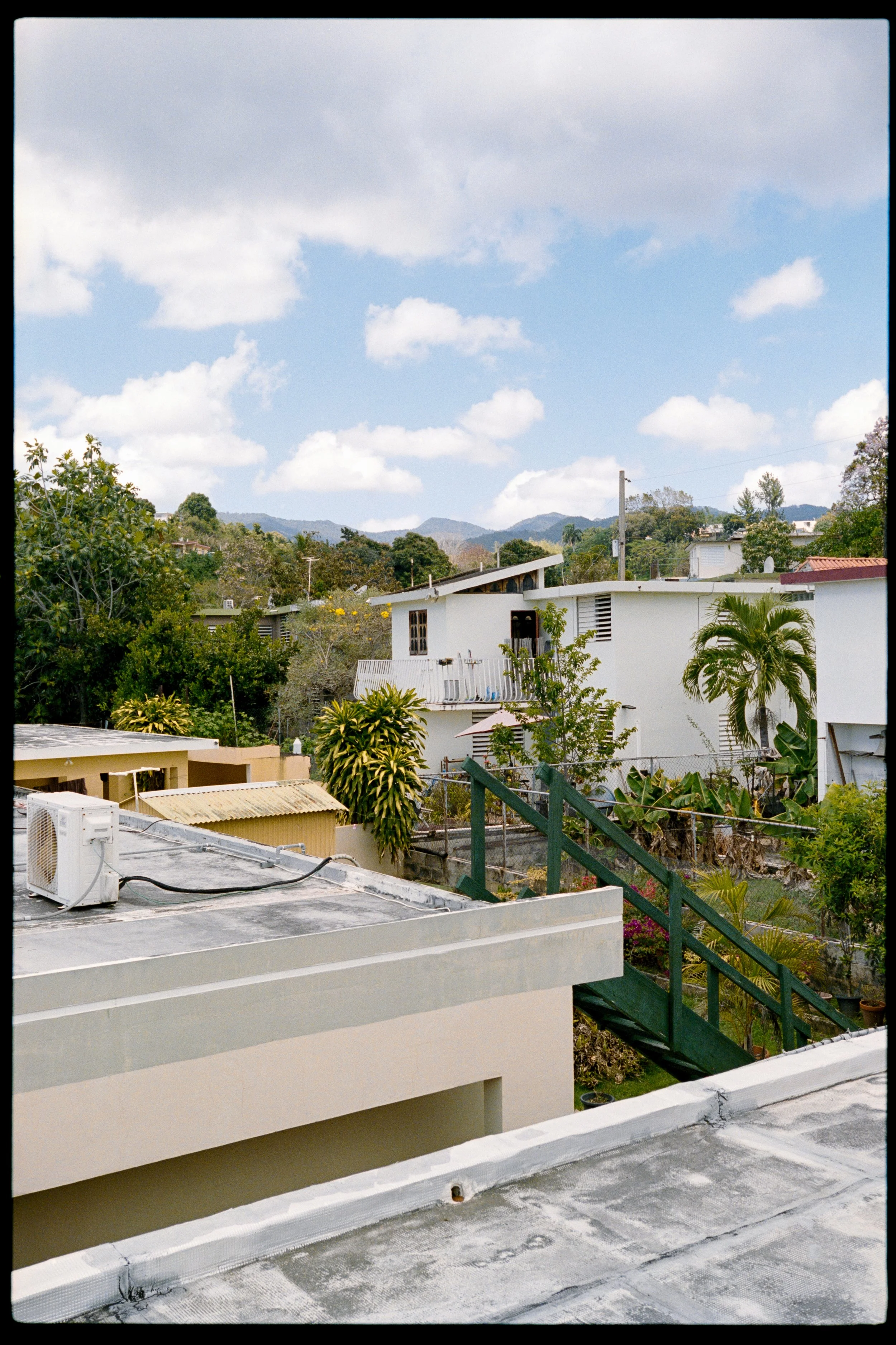 View of residential buildings with flat roofs, greenery, trees, and mountains in the background under a partly cloudy sky.