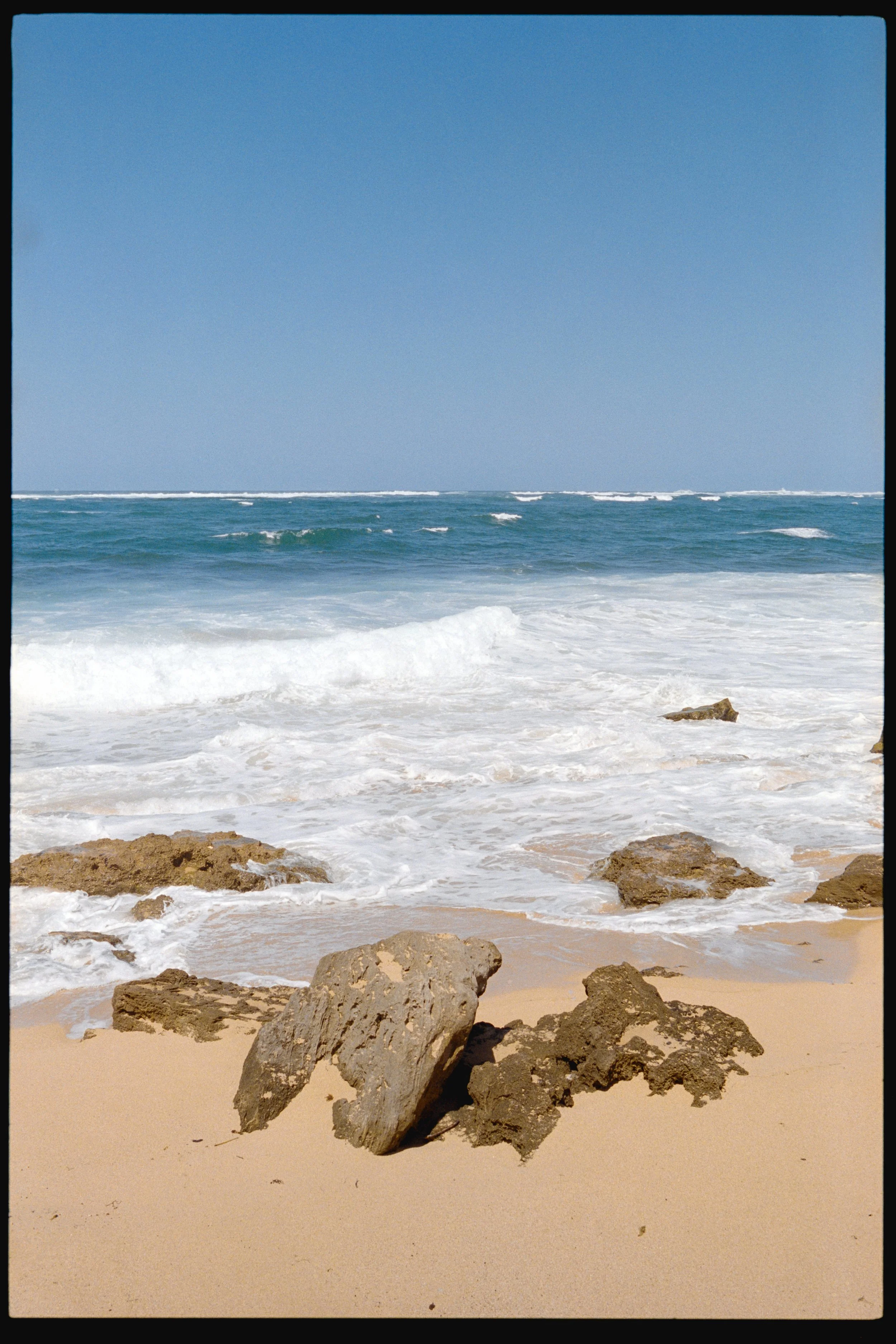 View of a sandy beach with rocks in the foreground, ocean waves, and a blue sky.