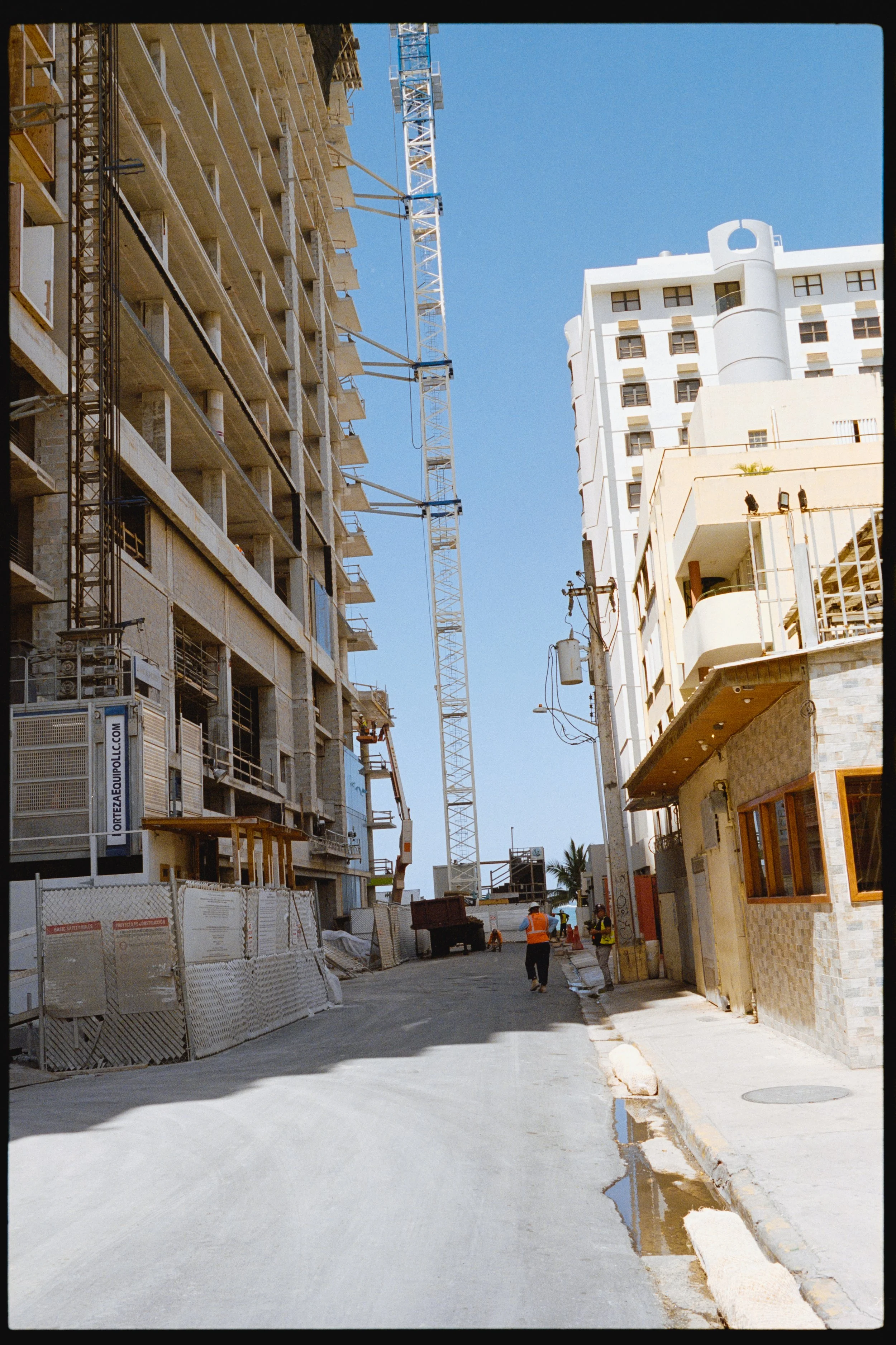A street under construction in an urban area with high-rise buildings, a crane, construction workers in orange vests, and construction barriers, with a bright blue sky overhead.