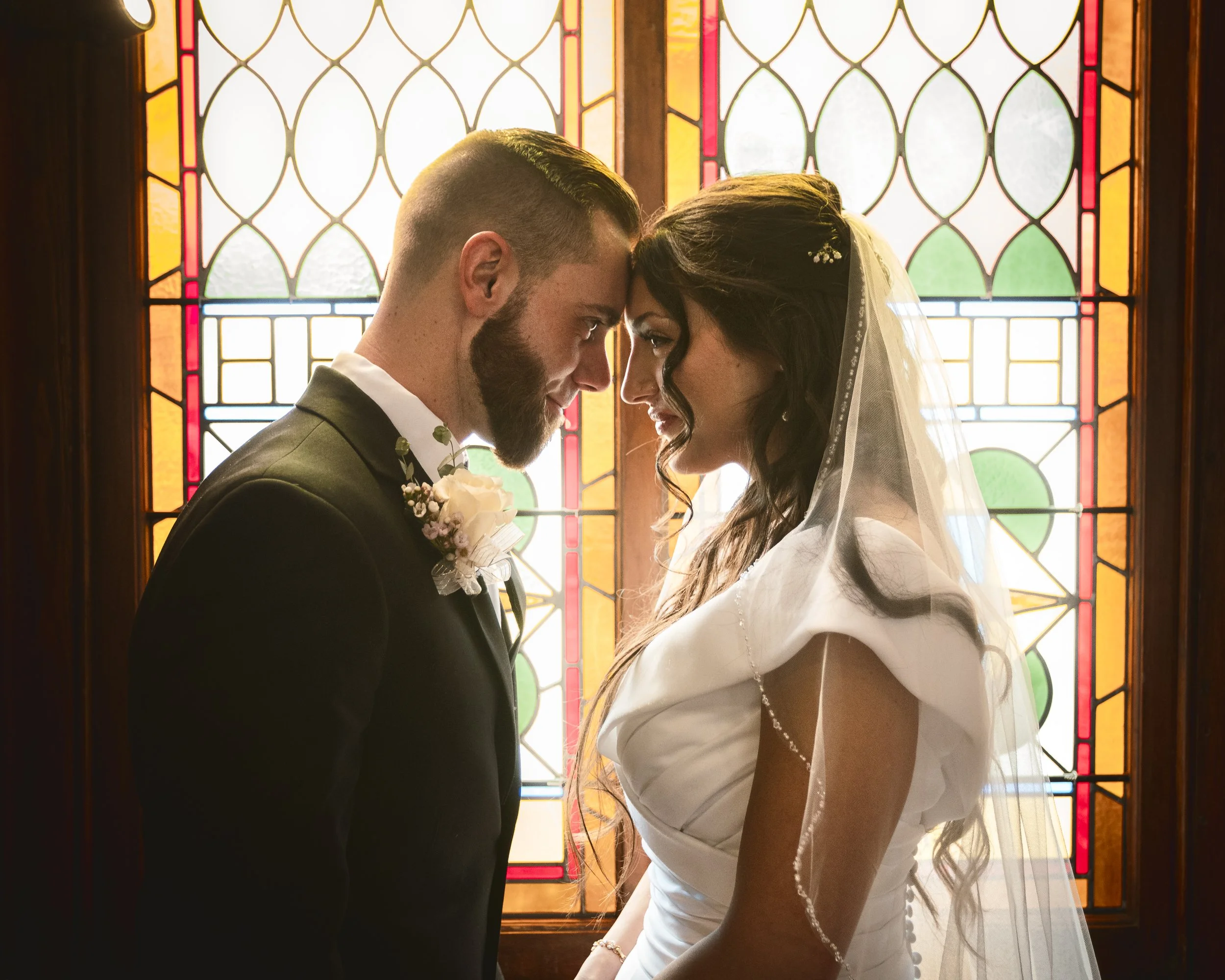 A bride and groom standing closely with their foreheads touching in front of stained glass windows during their wedding.