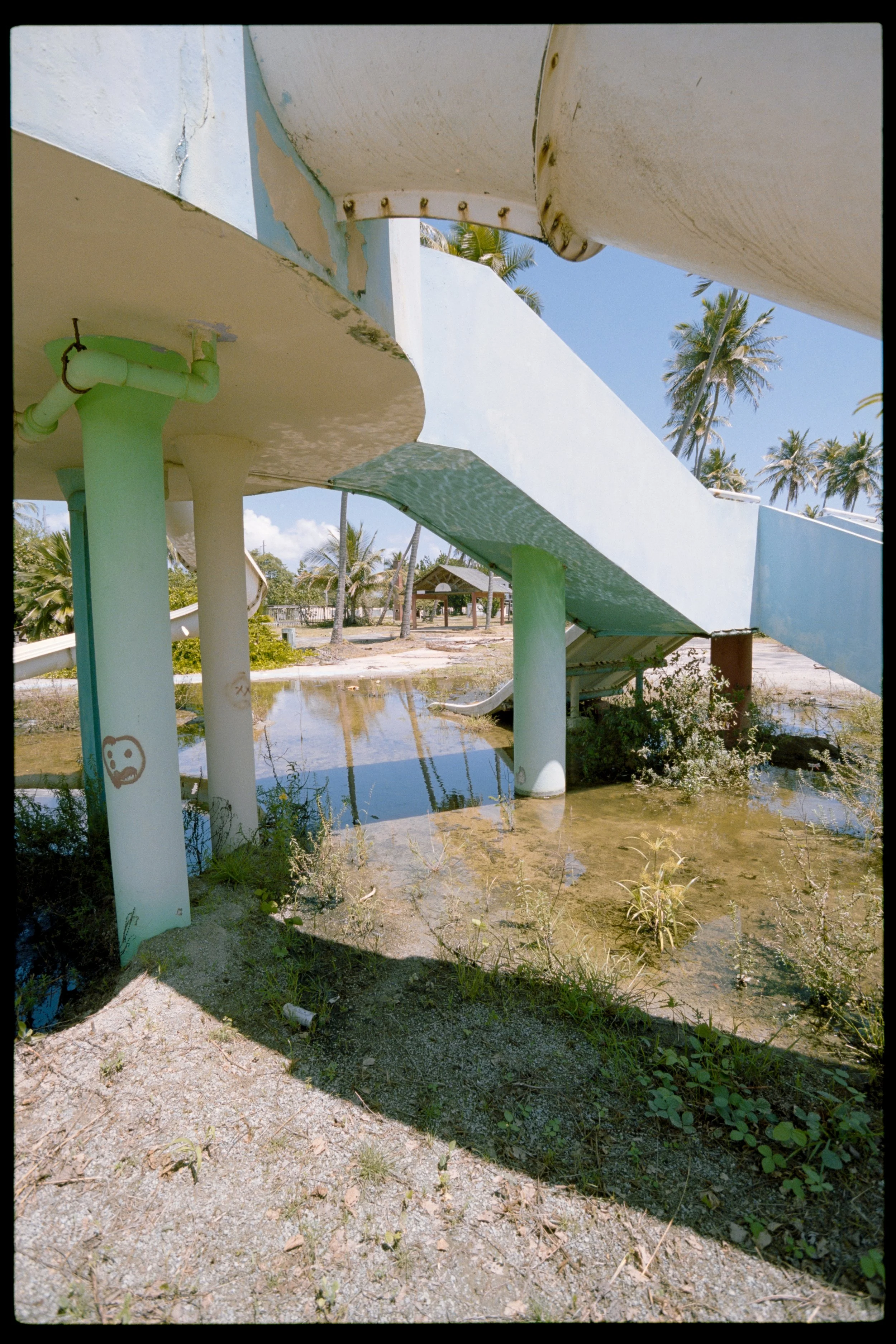 Empty water slide park with water remaining on the slide surface, overgrown plants, and trees in the background under a blue sky.