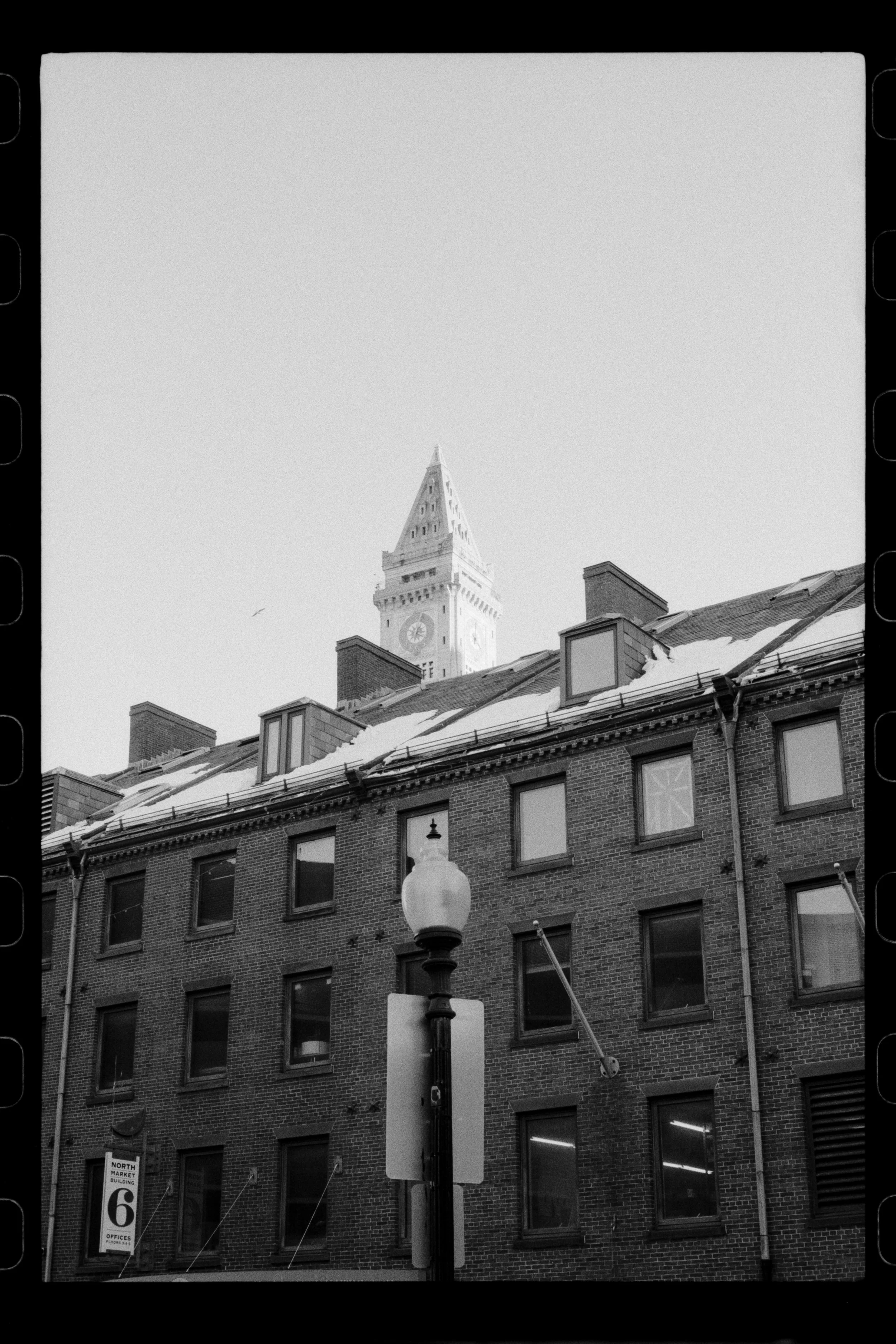 Black and white photo of a city street with a brick building, snow on the roof, a street lamp, and a clock tower in the background.