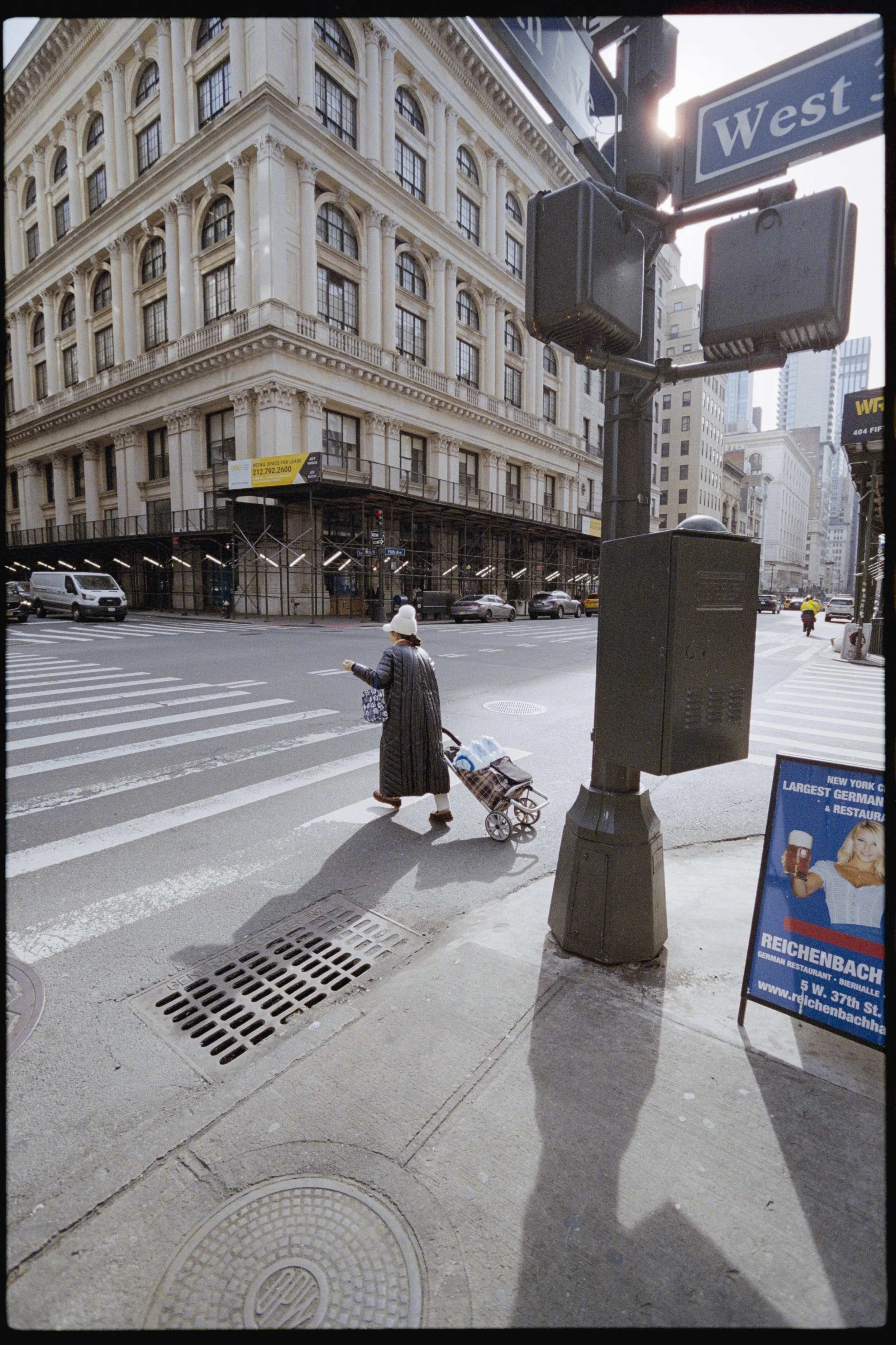 A person crossing a city street with a shopping cart, walking past a street sign that says 'West' and a black traffic signal box, in front of a historic building with scaffolding.
