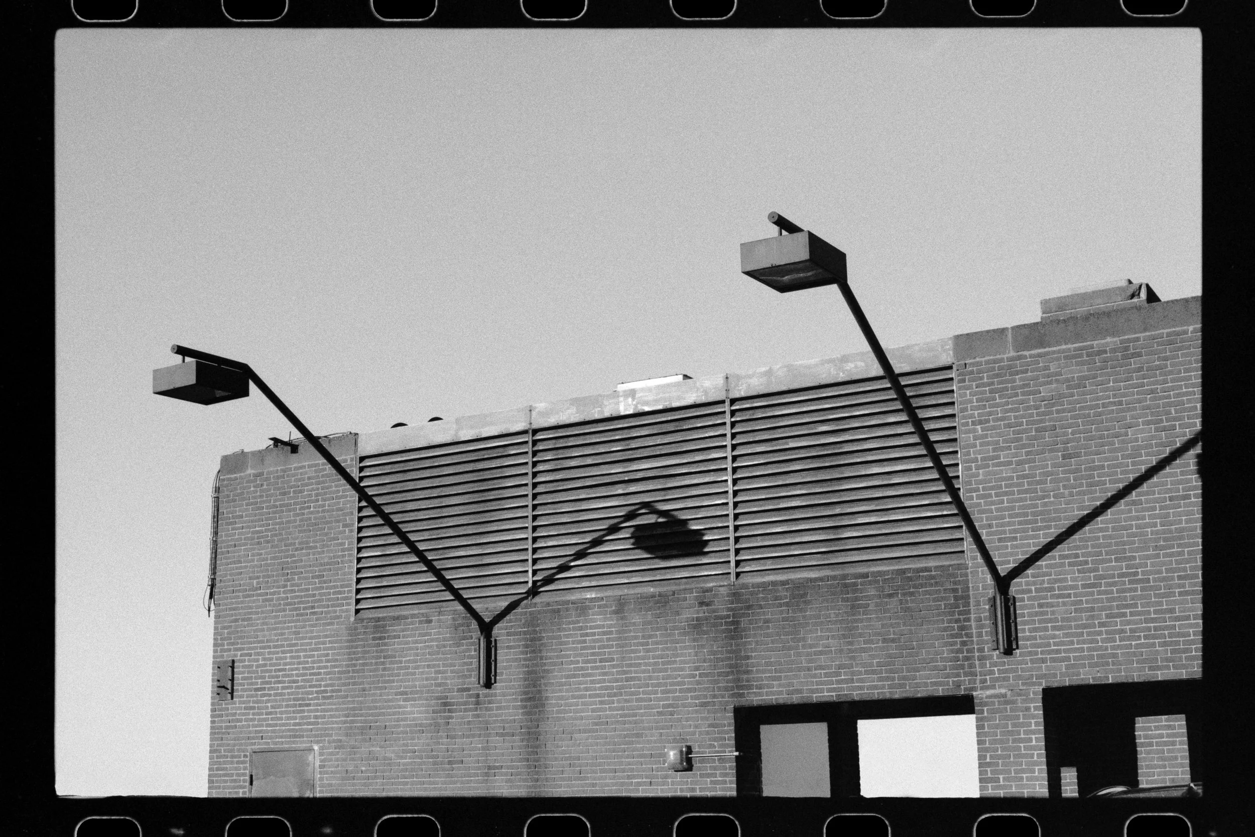 Black and white photo of a brick building with horizontal siding and scaffolding. Two streetlights are angled towards the building, casting shadows on the wall. The sky is clear and featureless.