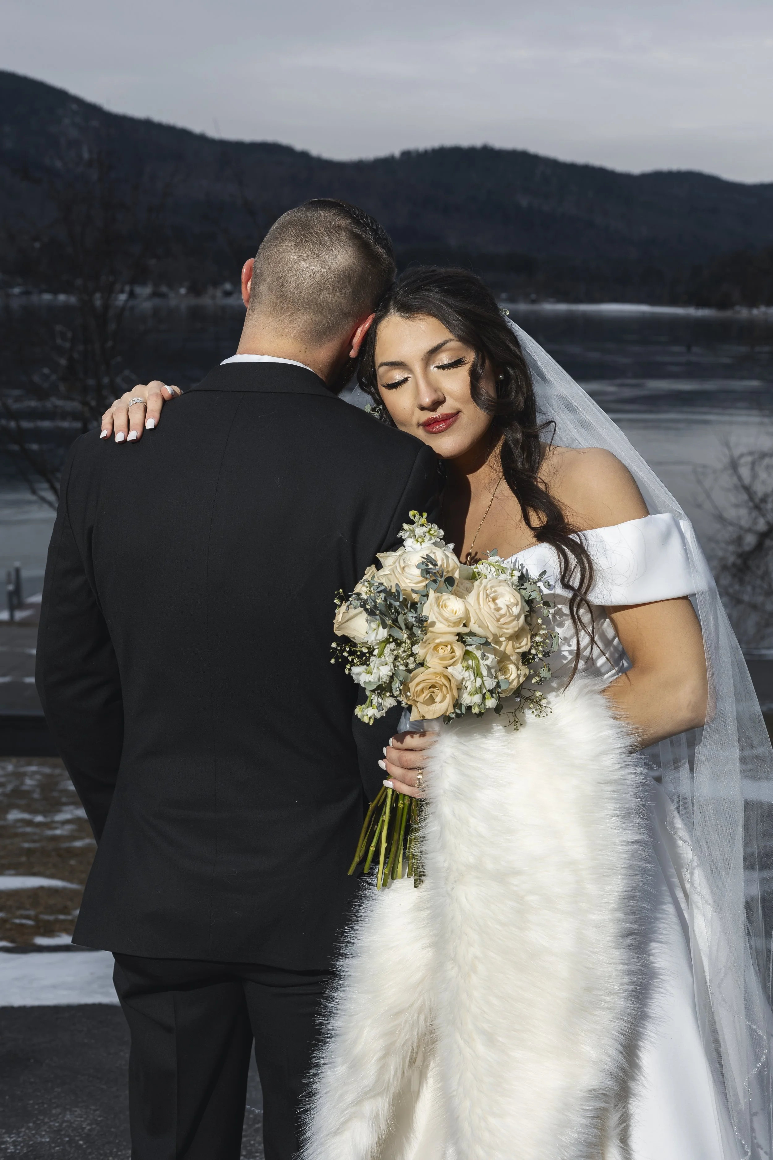 A bride and groom embrace outdoors near a river with mountains in the background. The bride holds a bouquet of white roses and wears a white off-shoulder wedding dress with a fur wrap. The groom wears a black suit.