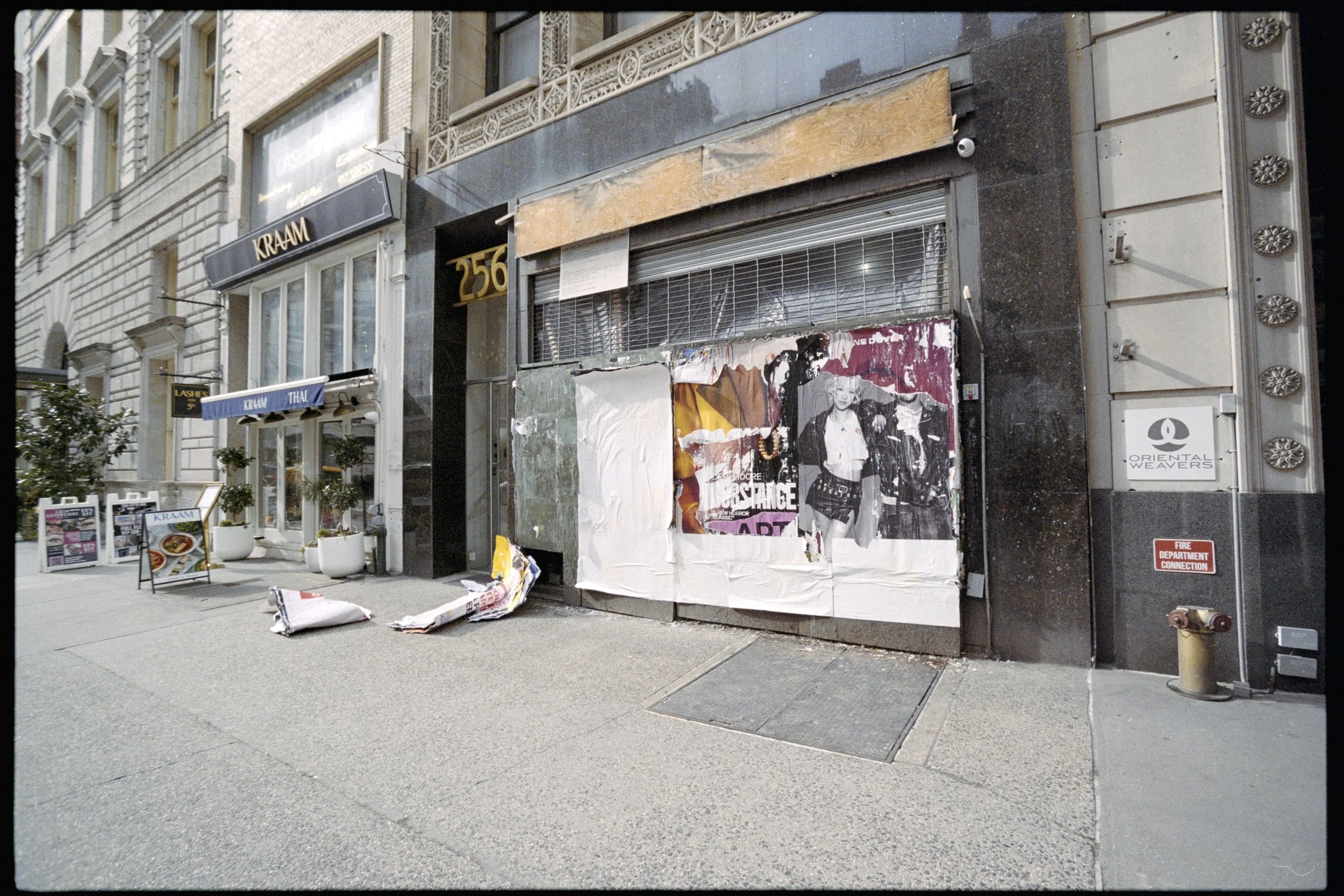 Empty storefront with boarded windows, torn posters, and scattered newspapers on sidewalk, with adjacent buildings and a fire department connection sign in urban setting.