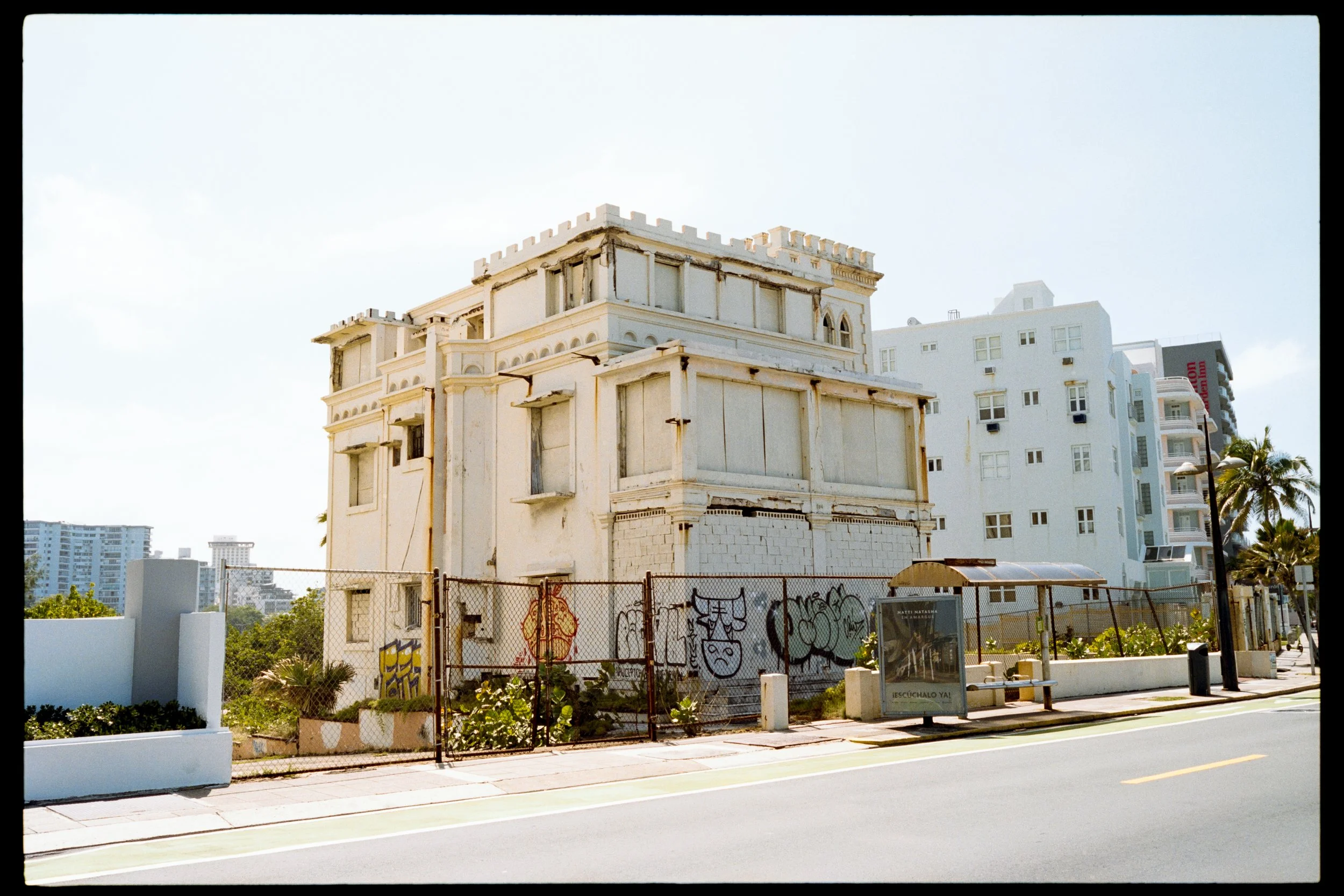 An unfinished white building with ornate architectural details next to modern high-rise buildings, surrounded by a chain-link fence with graffiti.