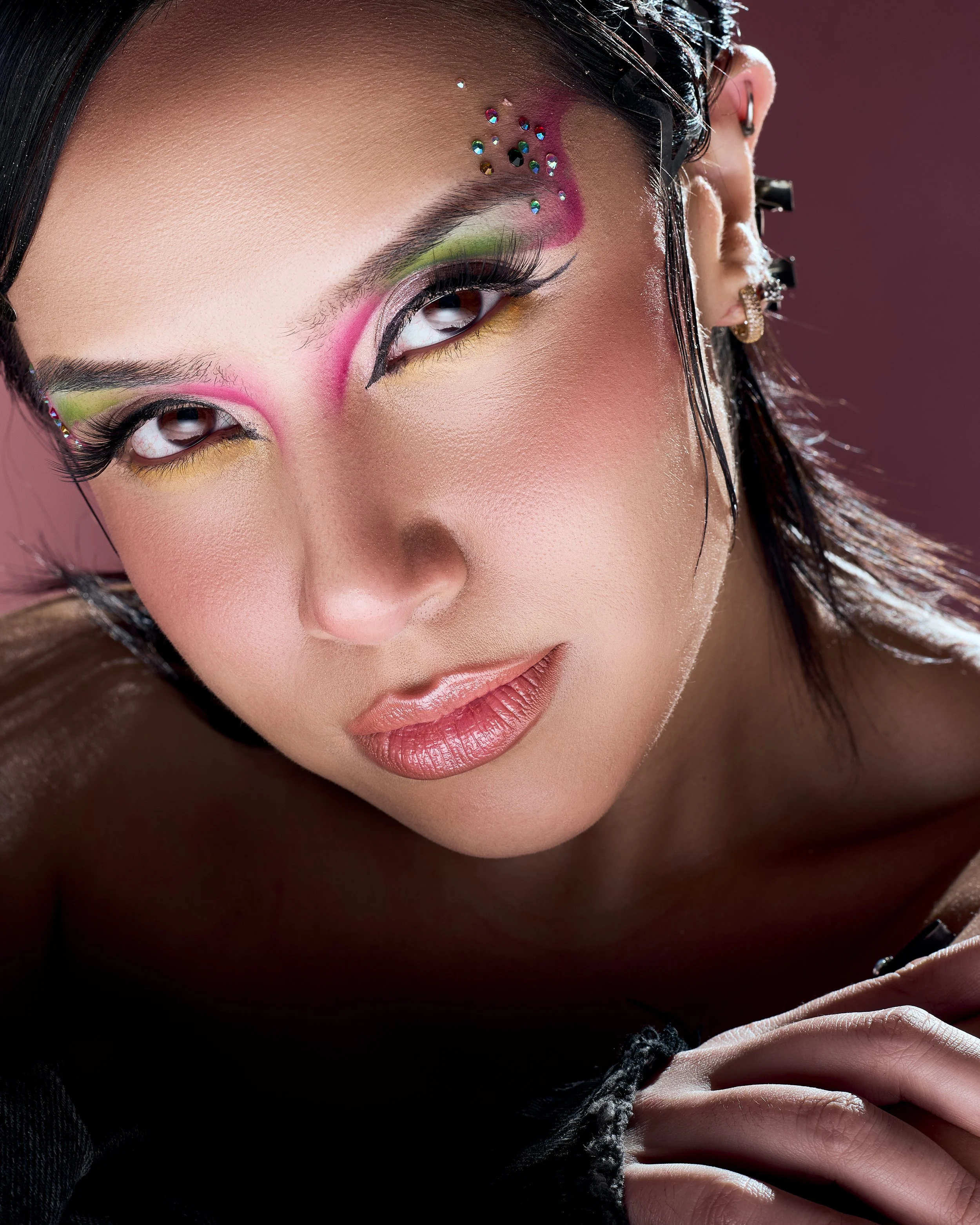 Close-up of a woman with colorful eye makeup, rhinestones on her eyelids, and pink lipstick, with wet hair and multiple earrings.