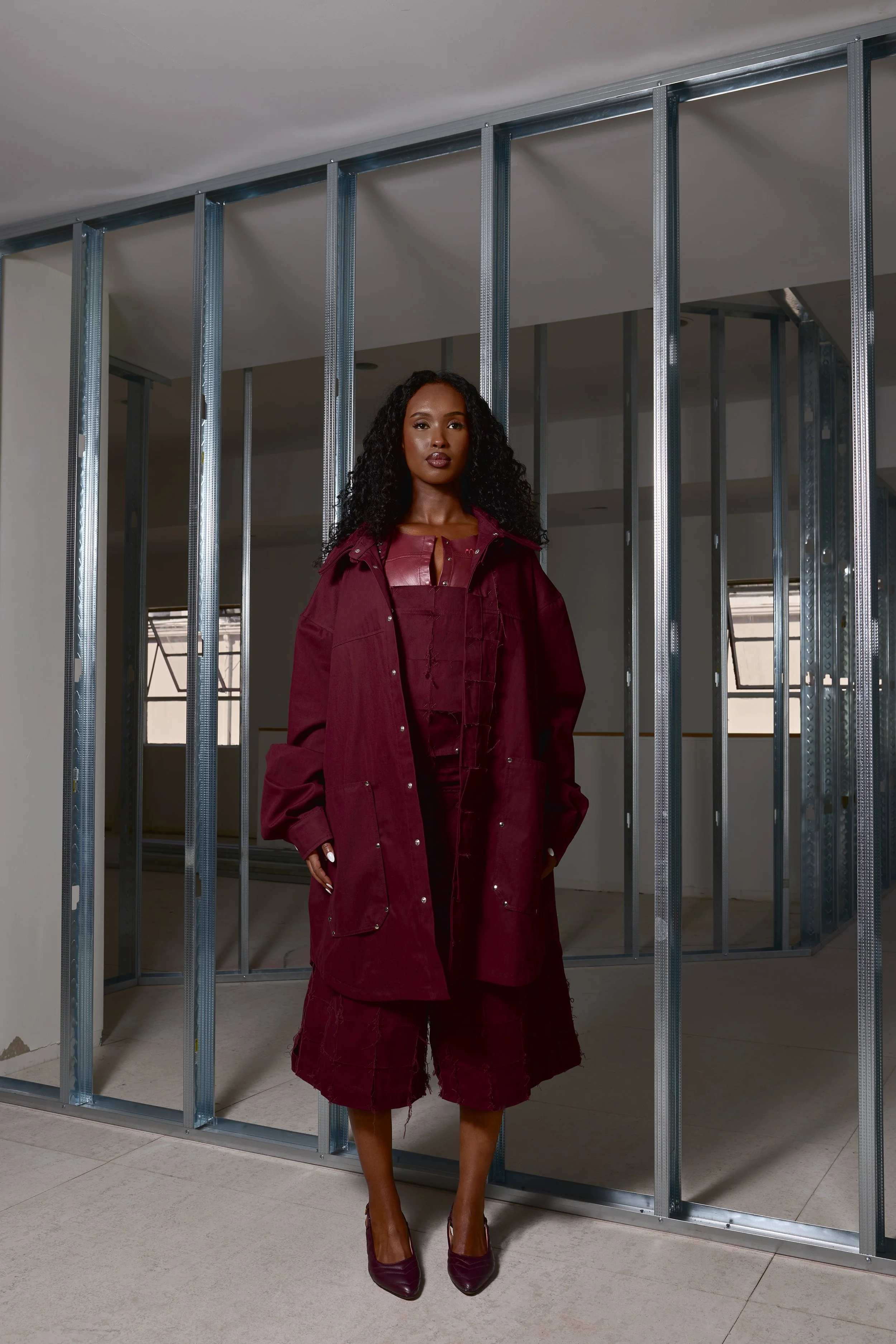 A woman in a burgundy outfit stands in front of an unfinished room with metal framing and windows.