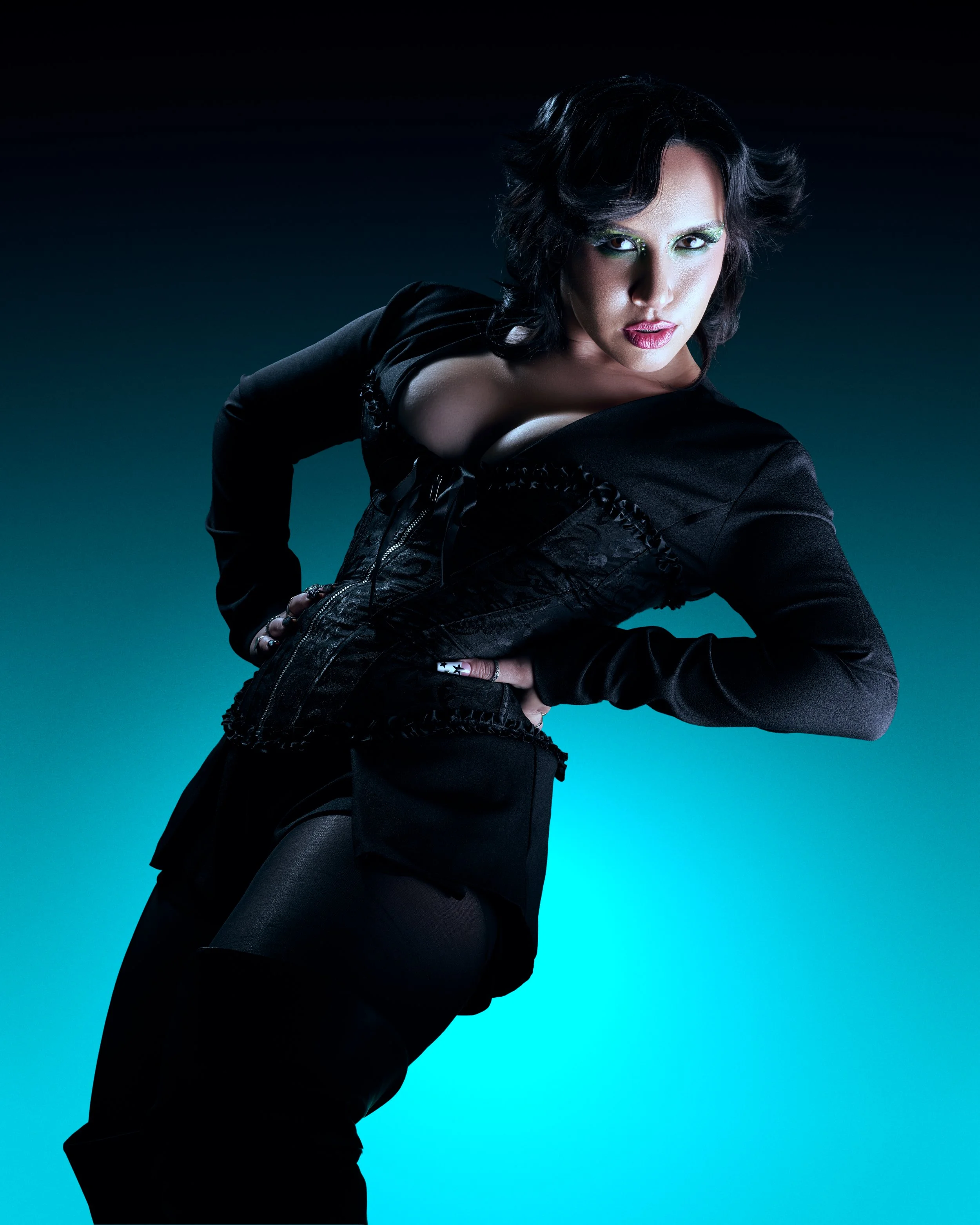 A woman with short dark hair wearing black clothing and bright makeup poses against a gradient background.