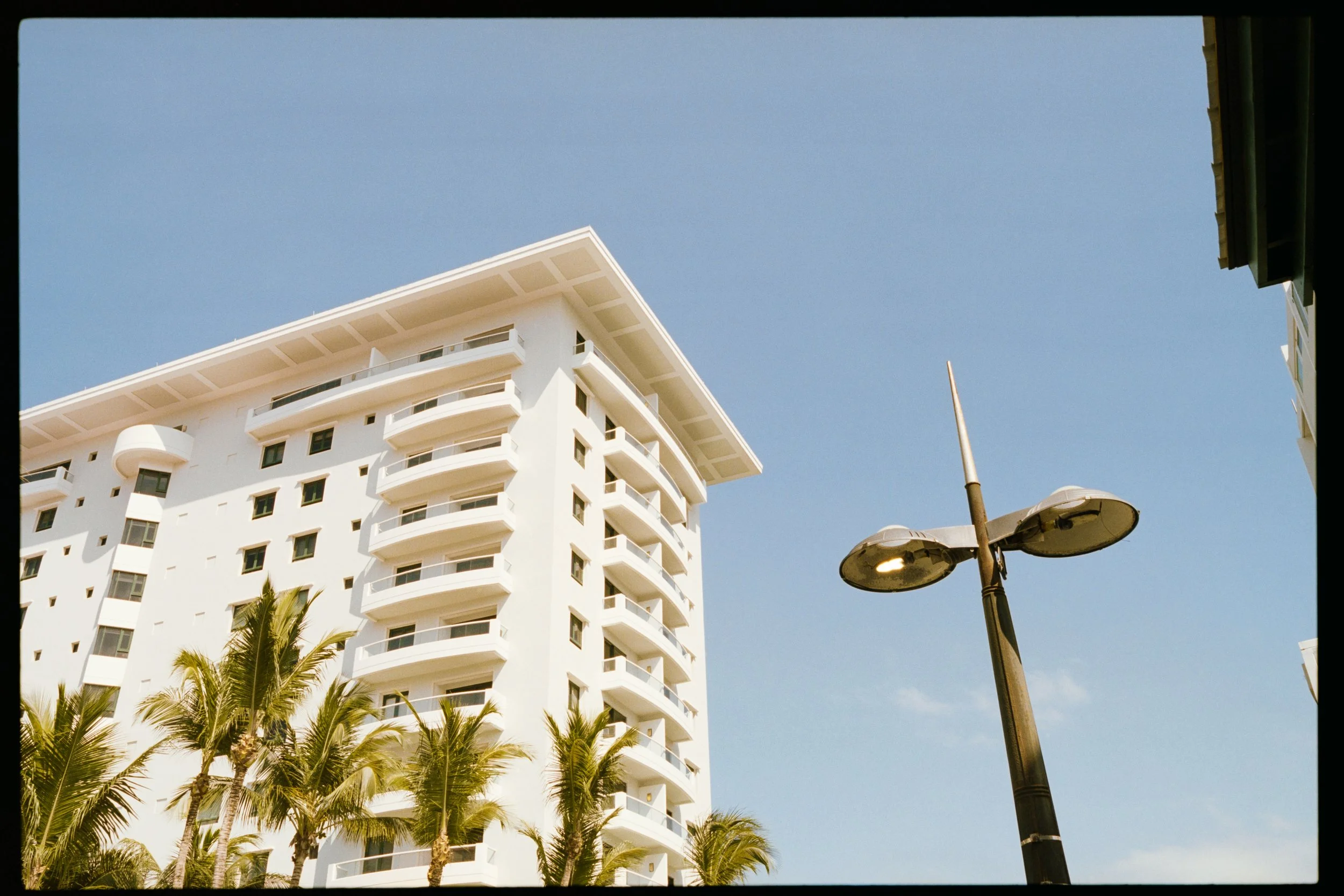 A white multi-story building with curved balconies, palm trees in front, a streetlamp, and a clear blue sky.