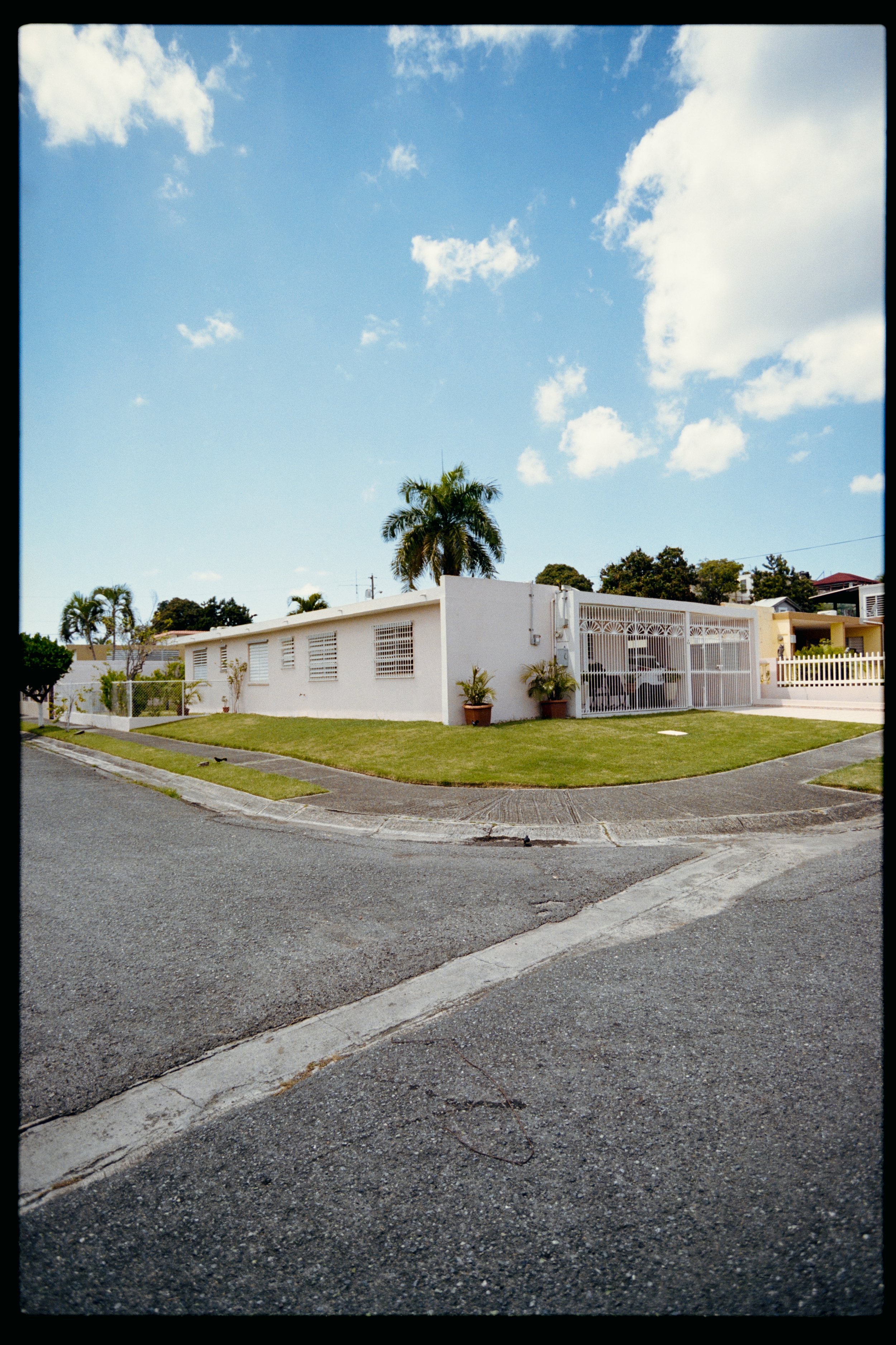 A residential street corner with a white house, palm trees, and a neatly maintained lawn under a blue sky with clouds.